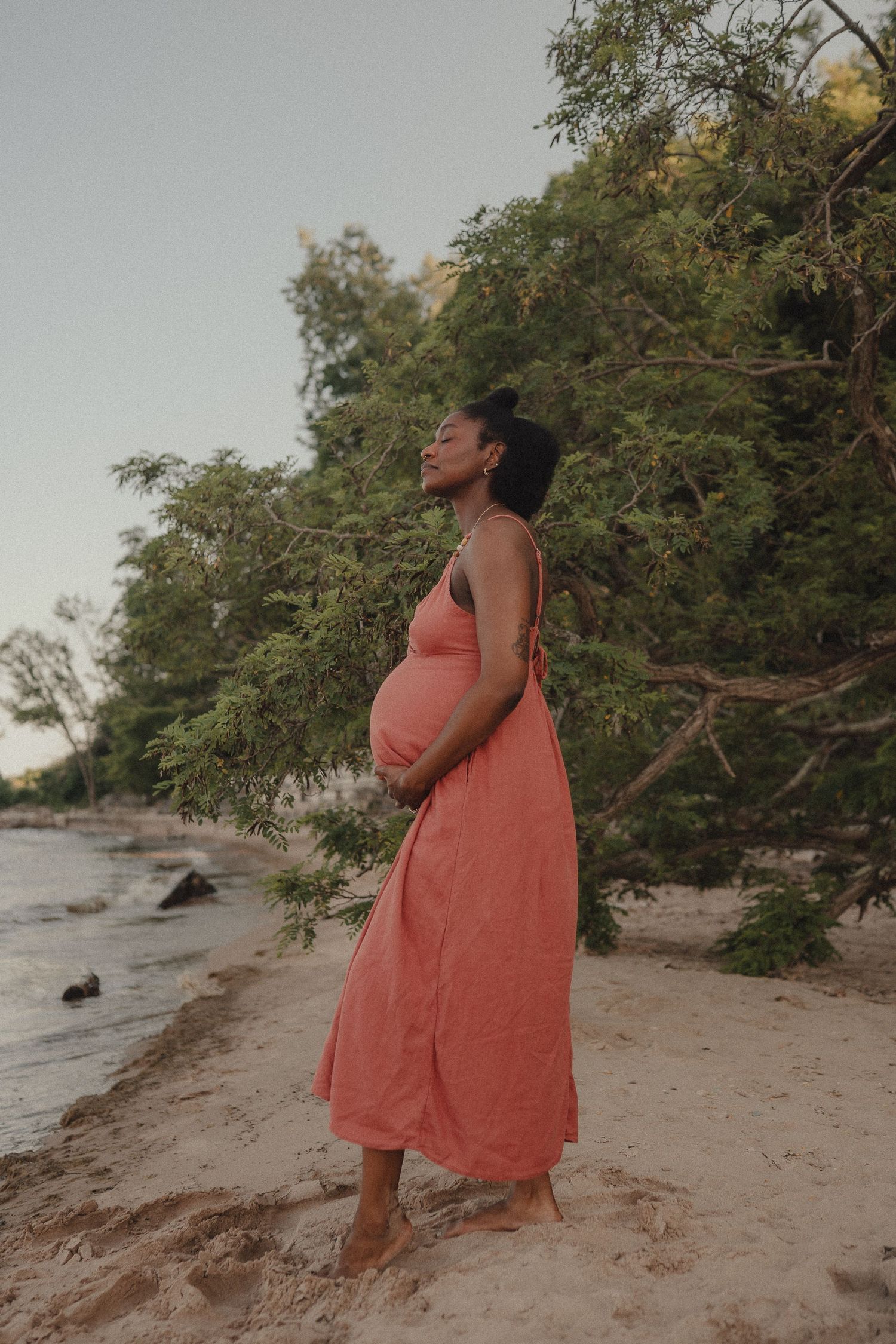 A serene pregnant Black woman in coral dress stands barefoot on a sandy beach among green trees.