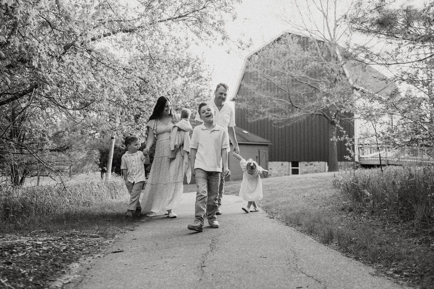 Black and white photo of a family walking down a tree-lined path near a rustic barn.