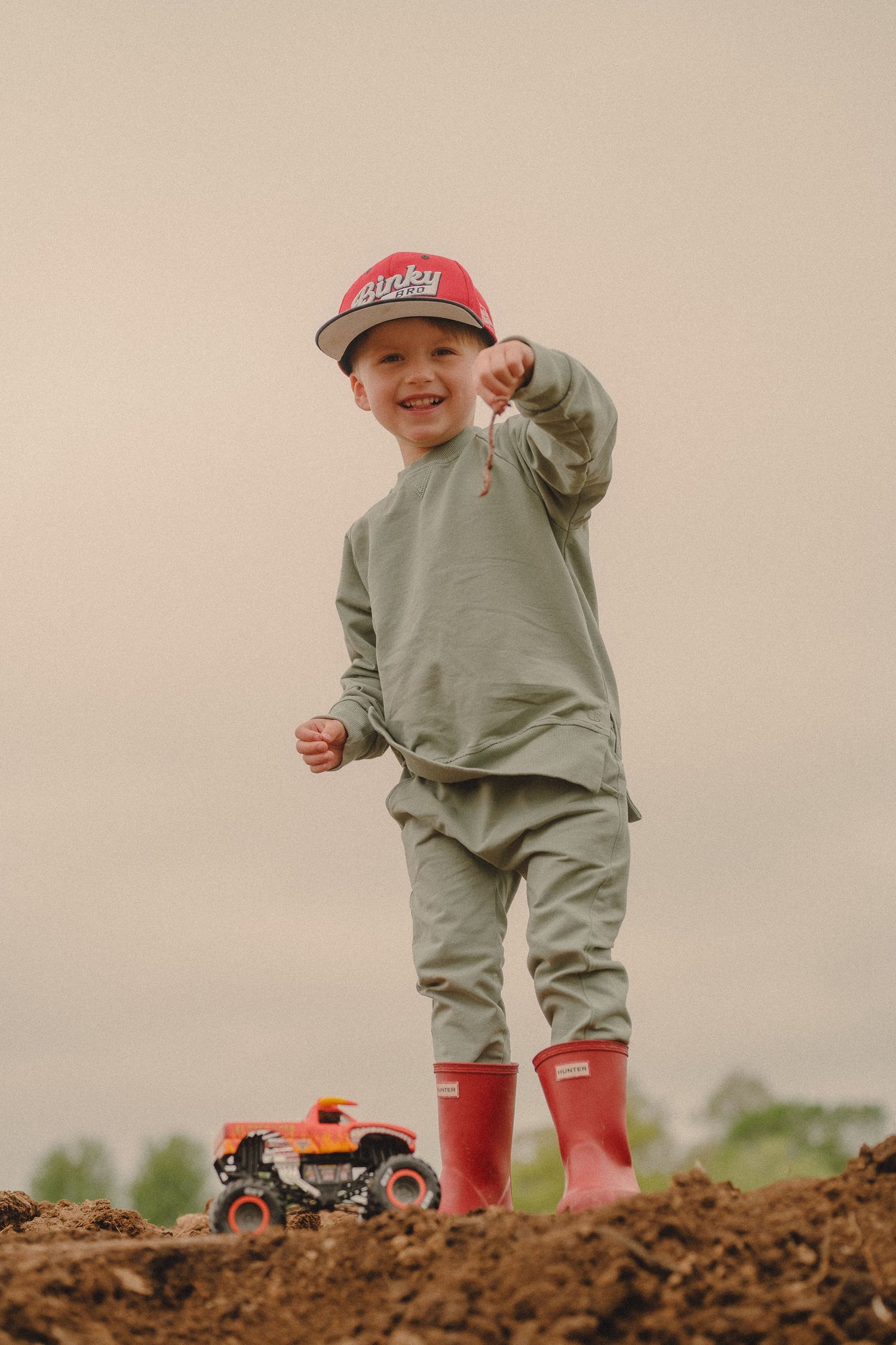 Happy young boy standing on dirt mound with Monster Truck toy wearing red cap and boots.