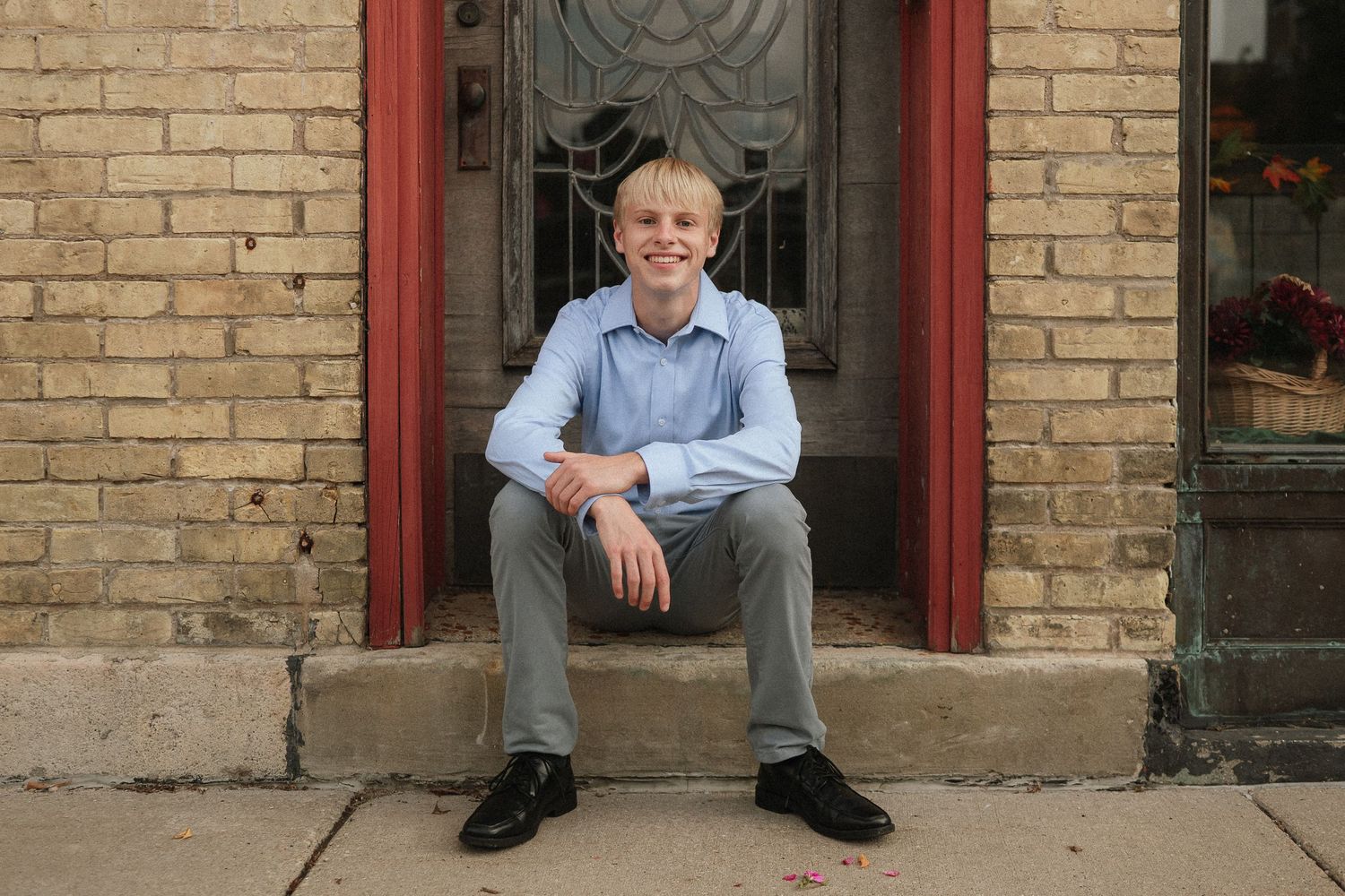 Someone sits on the front steps of a brick building with a decorative red door frame, smiling at the camera.