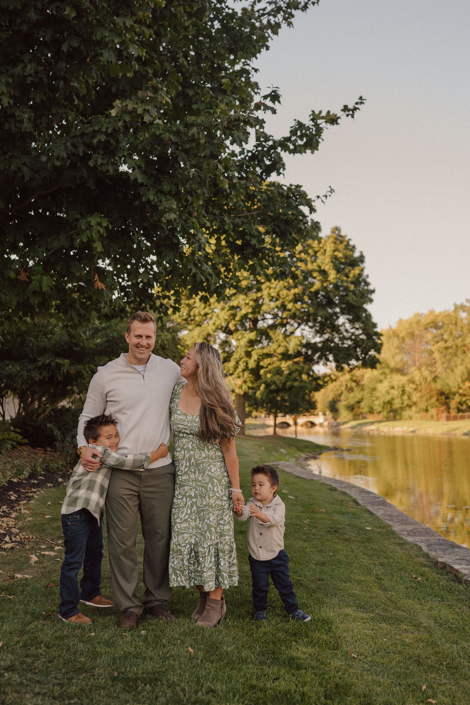 A family enjoys a warm evening together by a tranquil lake with trees and grass along the shoreline.