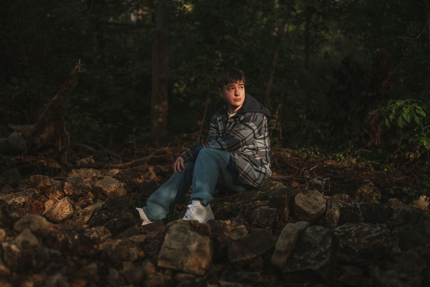 Person in blue jacket sits on rustic stone wall surrounded by autumn leaves in dark wooded setting.