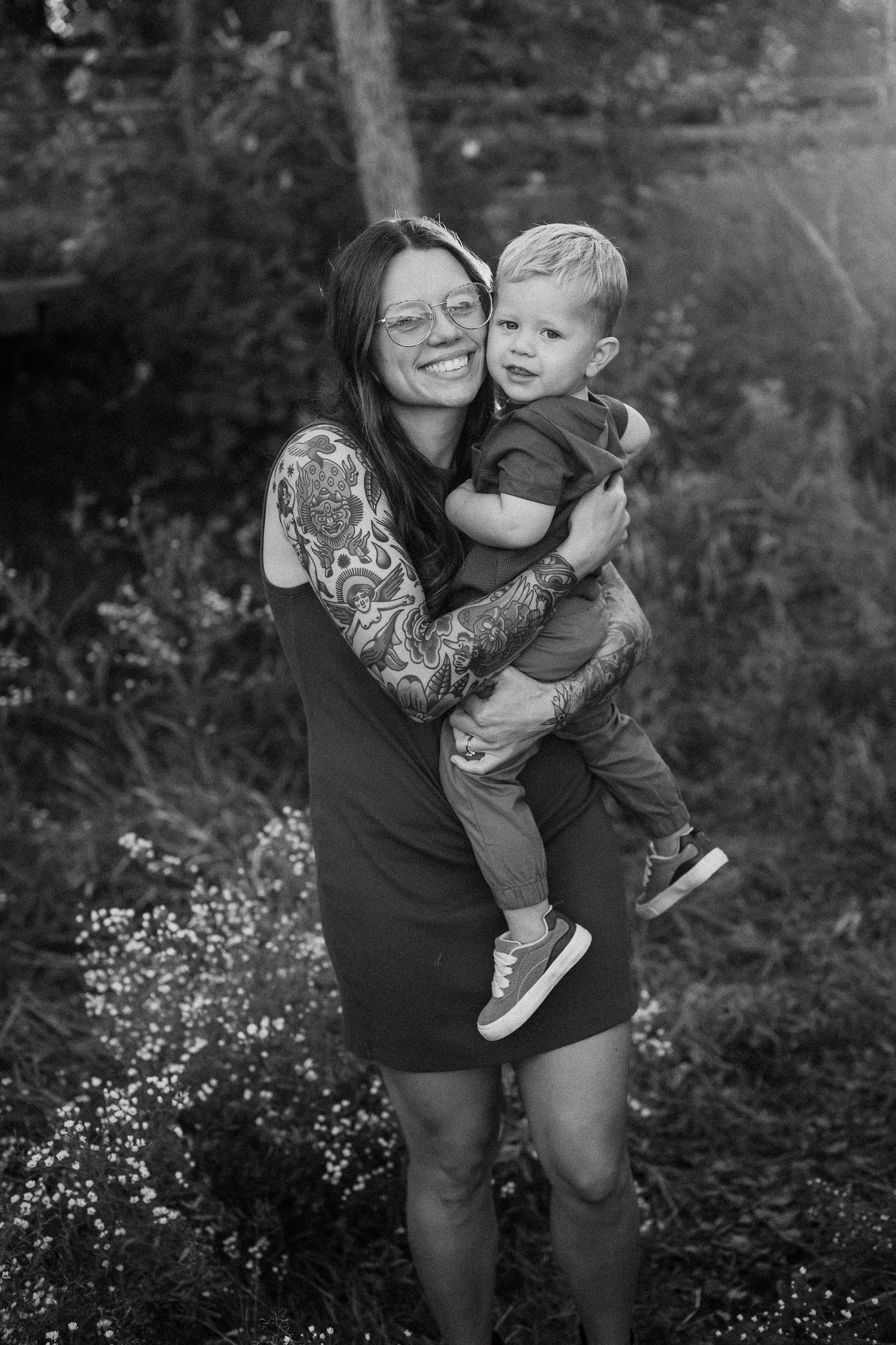 A black and white portrait of a woman with sleeve tattoos holding a small child outdoors in a natural setting.