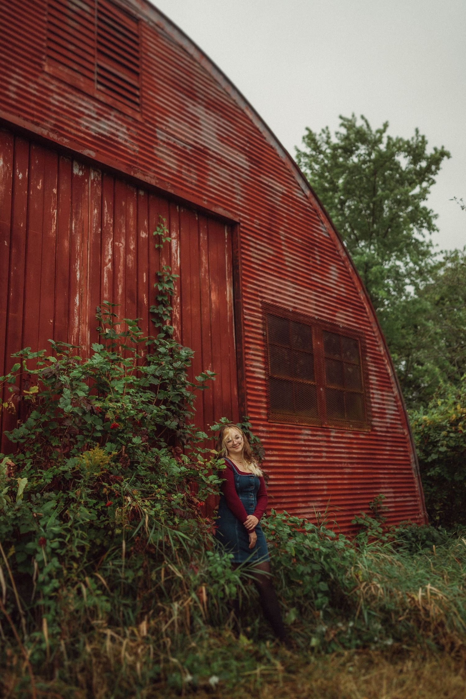 A rustic portrait against a large red barn with curved metal roofing surrounded by wild plants.