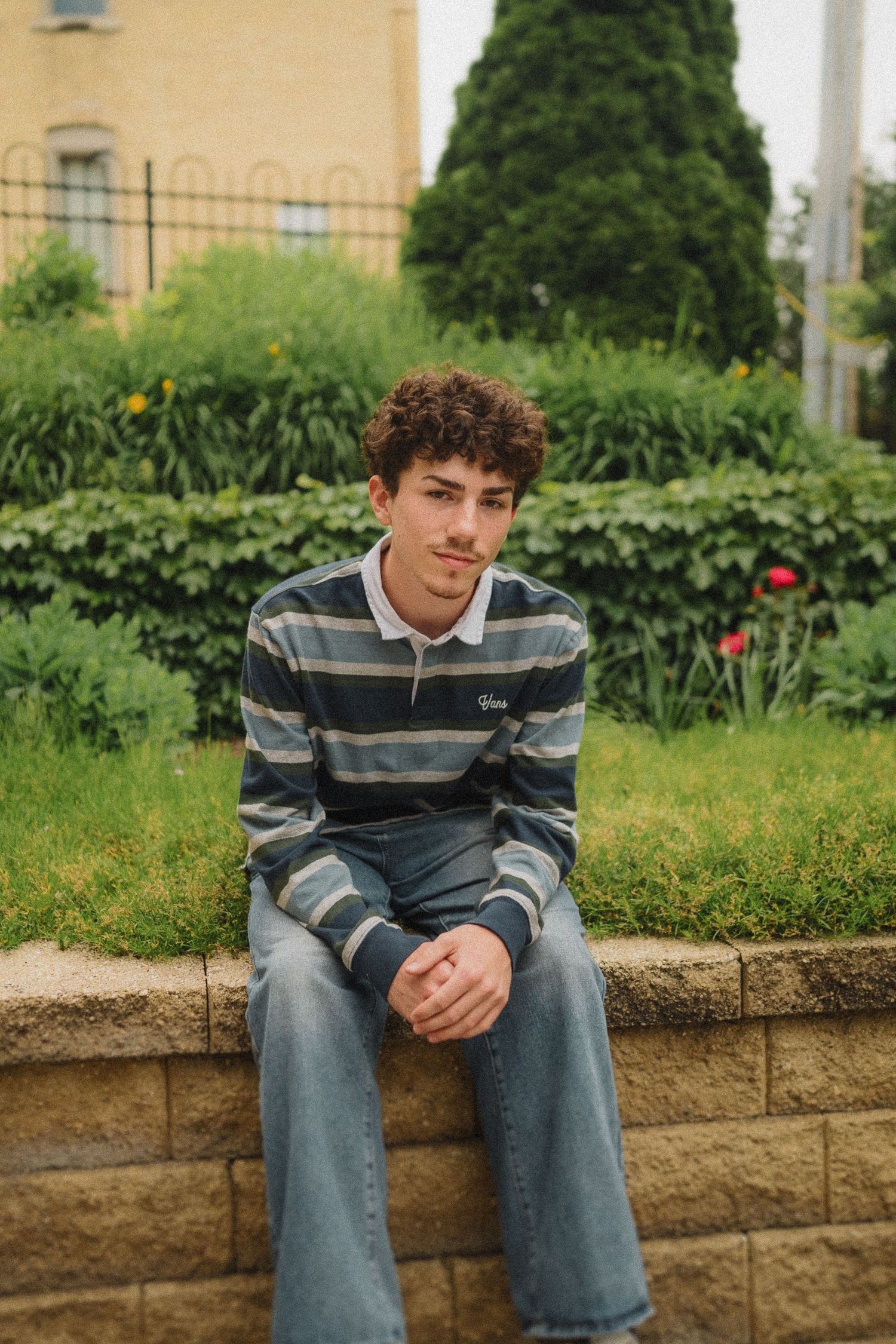 Casual portrait of someone in striped rugby shirt seated on stone wall with garden and building backdrop.