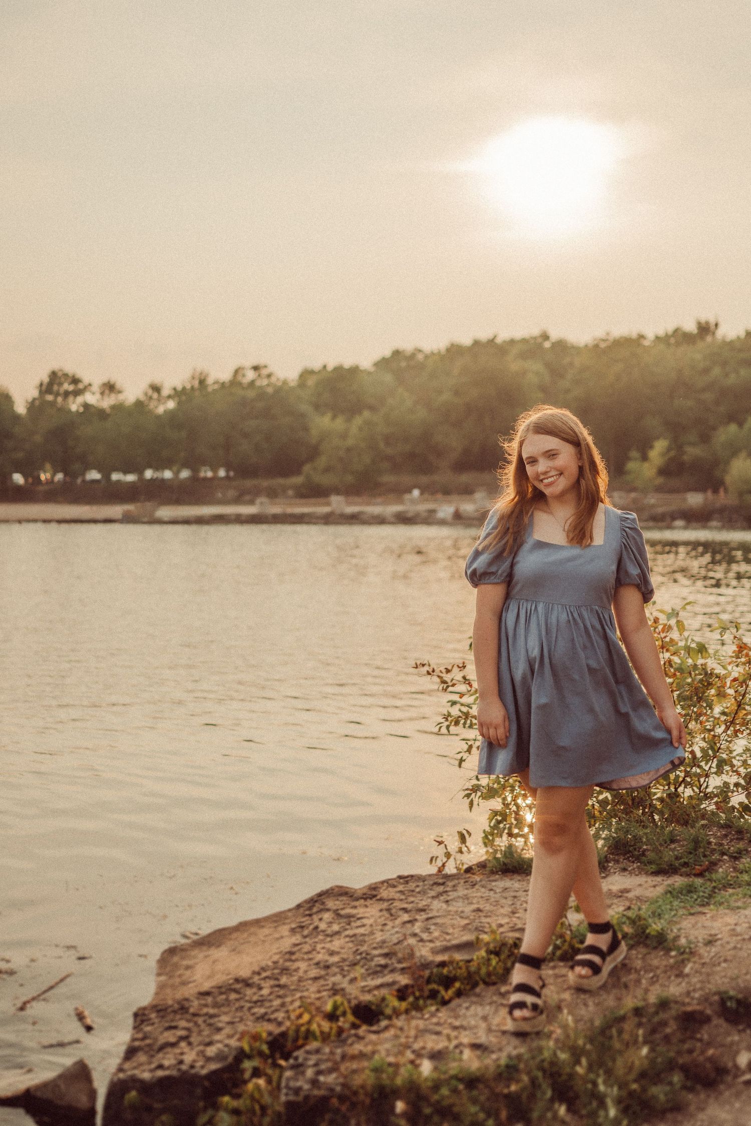 A sunset portrait by a lake featuring someone in a casual blue dress standing on rocky terrain.