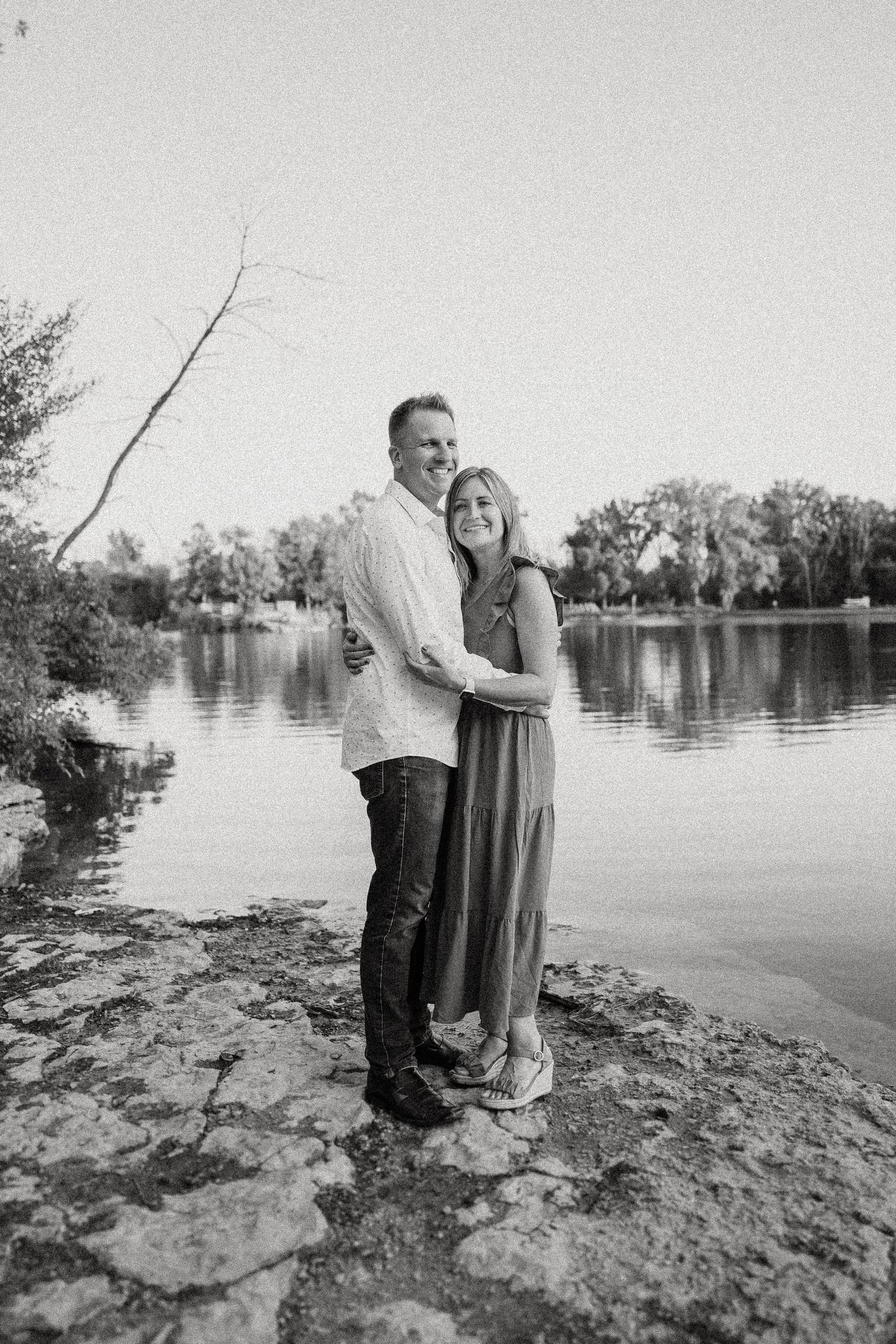 Black and white portrait of a couple embracing by a tranquil lake.