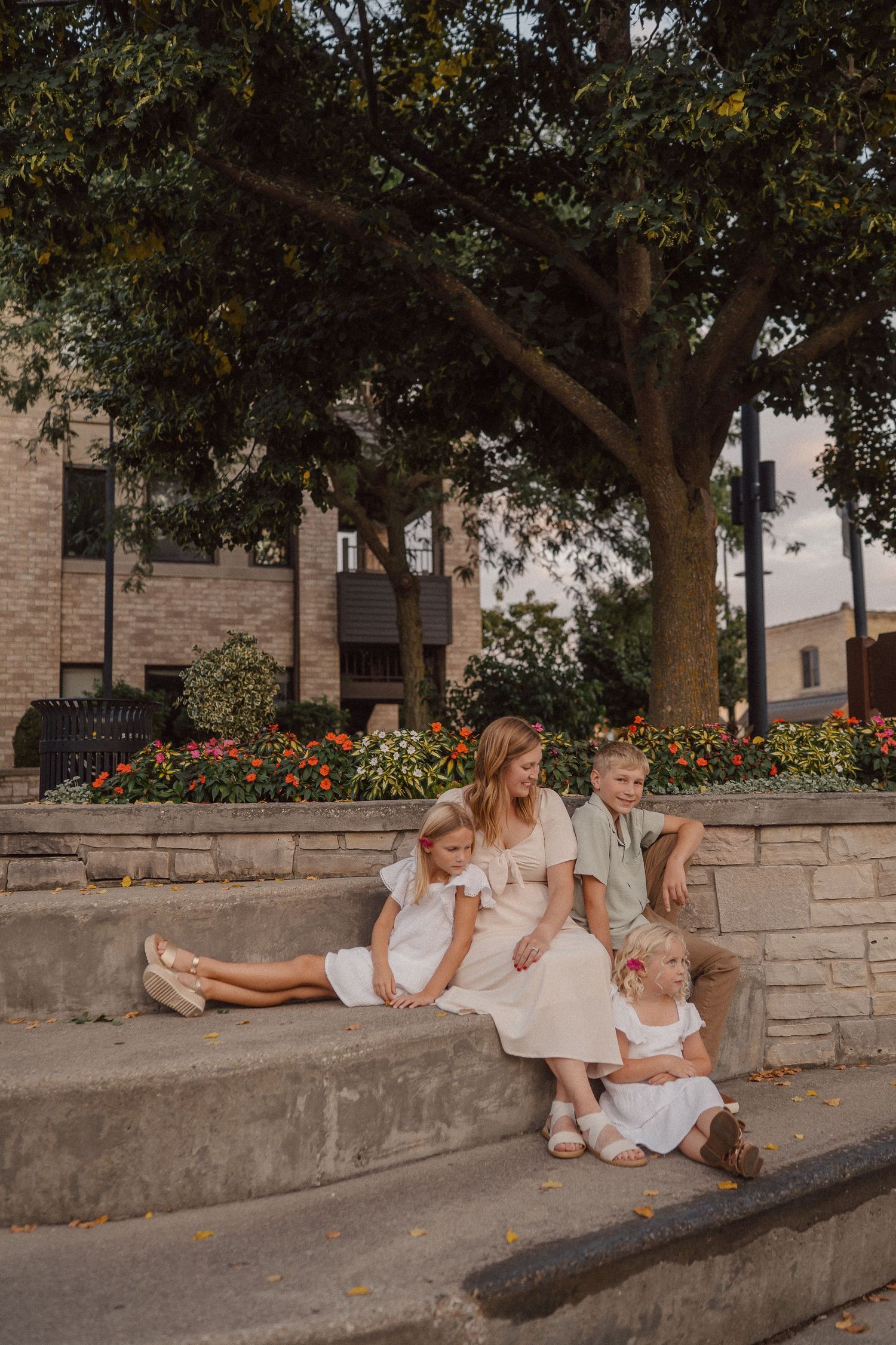 People in light colored outfits sit casually along a stone wall under shady trees.