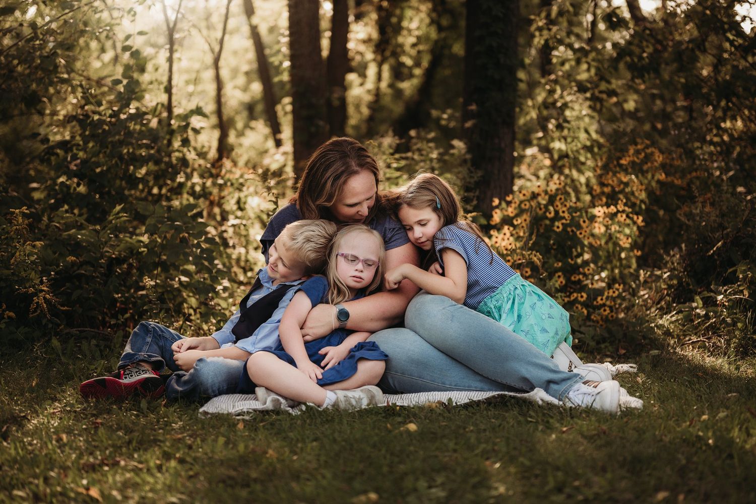 A loving family cuddles together while sitting on a blanket in a sunlit forest clearing.