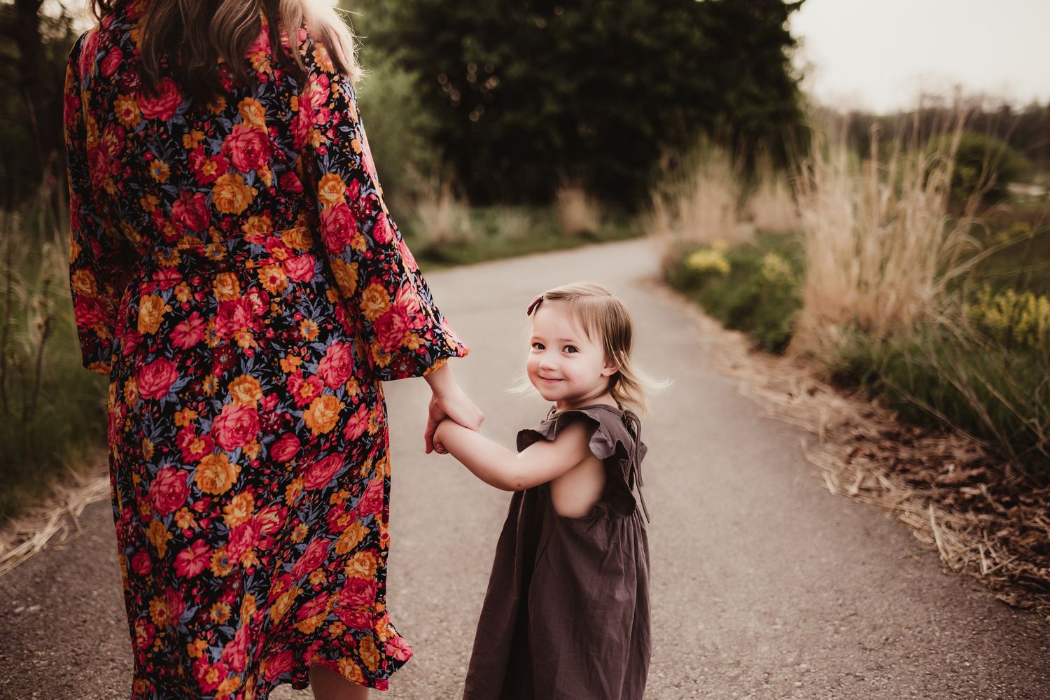 A colorful floral dress contrasts with a brown party dress on a path surrounded by natural foliage.