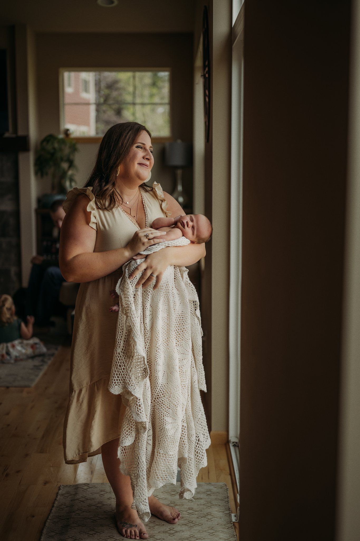 Woman in cream dress stands by window holding newborn while smiling in warm natural light.