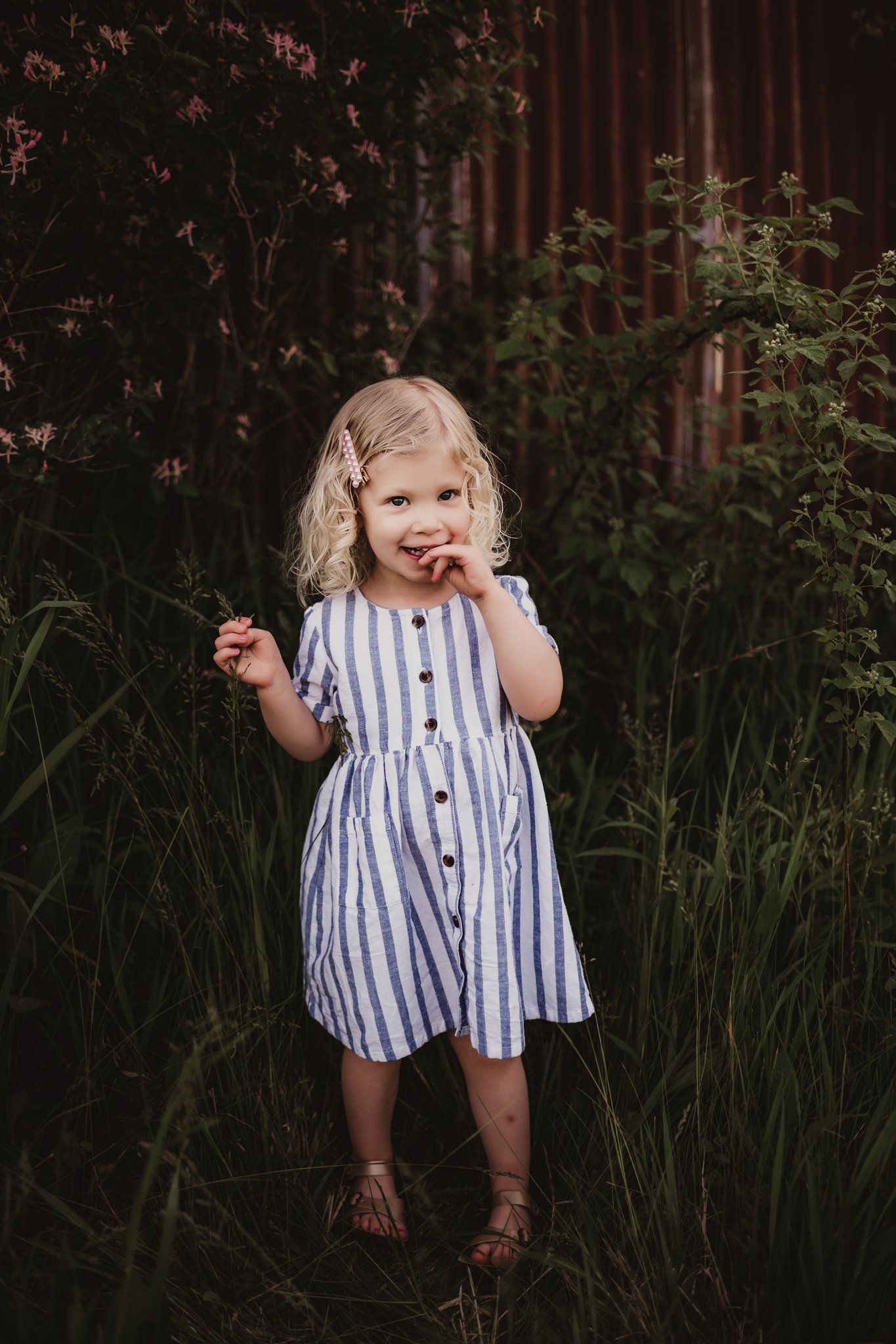 A playful child in a striped dress stands among wild grasses near a rustic building.