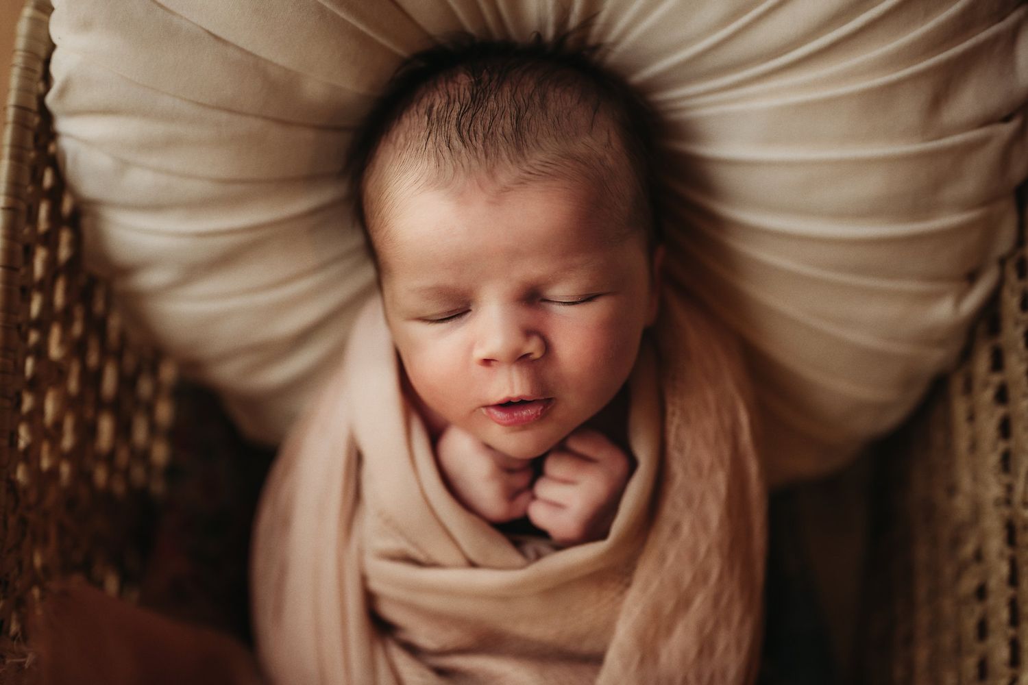 Close-up of sleeping newborn baby swaddled in beige fabric against cream pillow.