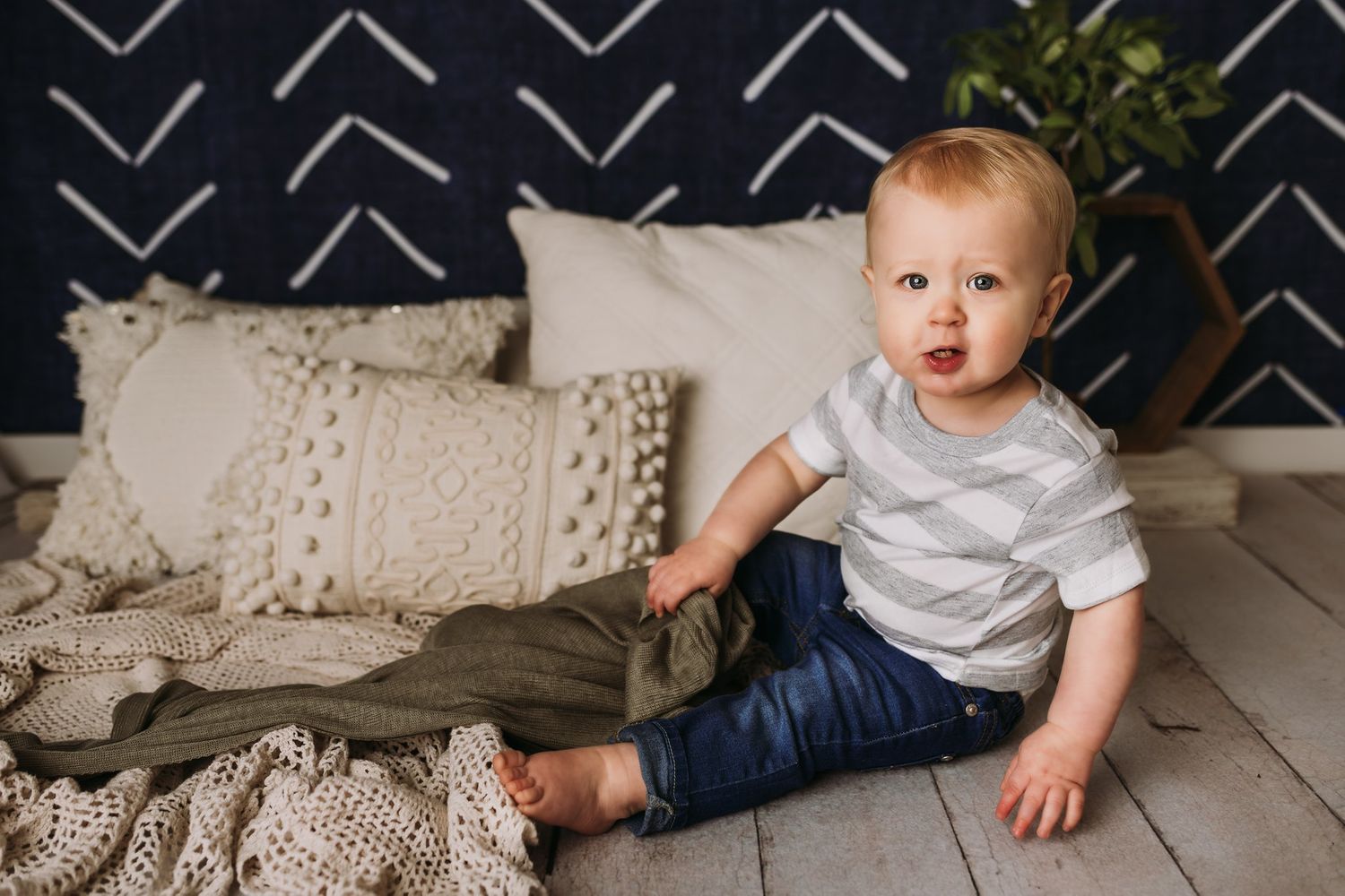 A toddler in casual wear sits on wooden flooring with decorative pillows and blankets.
