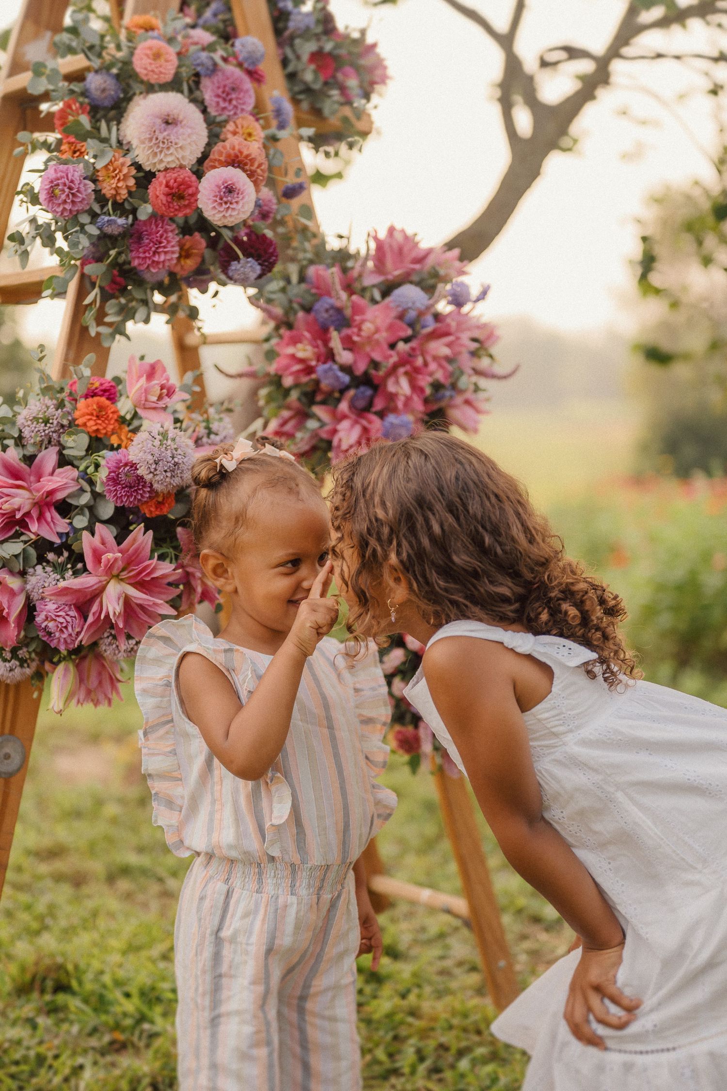 Sweet interaction between children near a wooden easel decorated with pink and purple flowers in a garden.