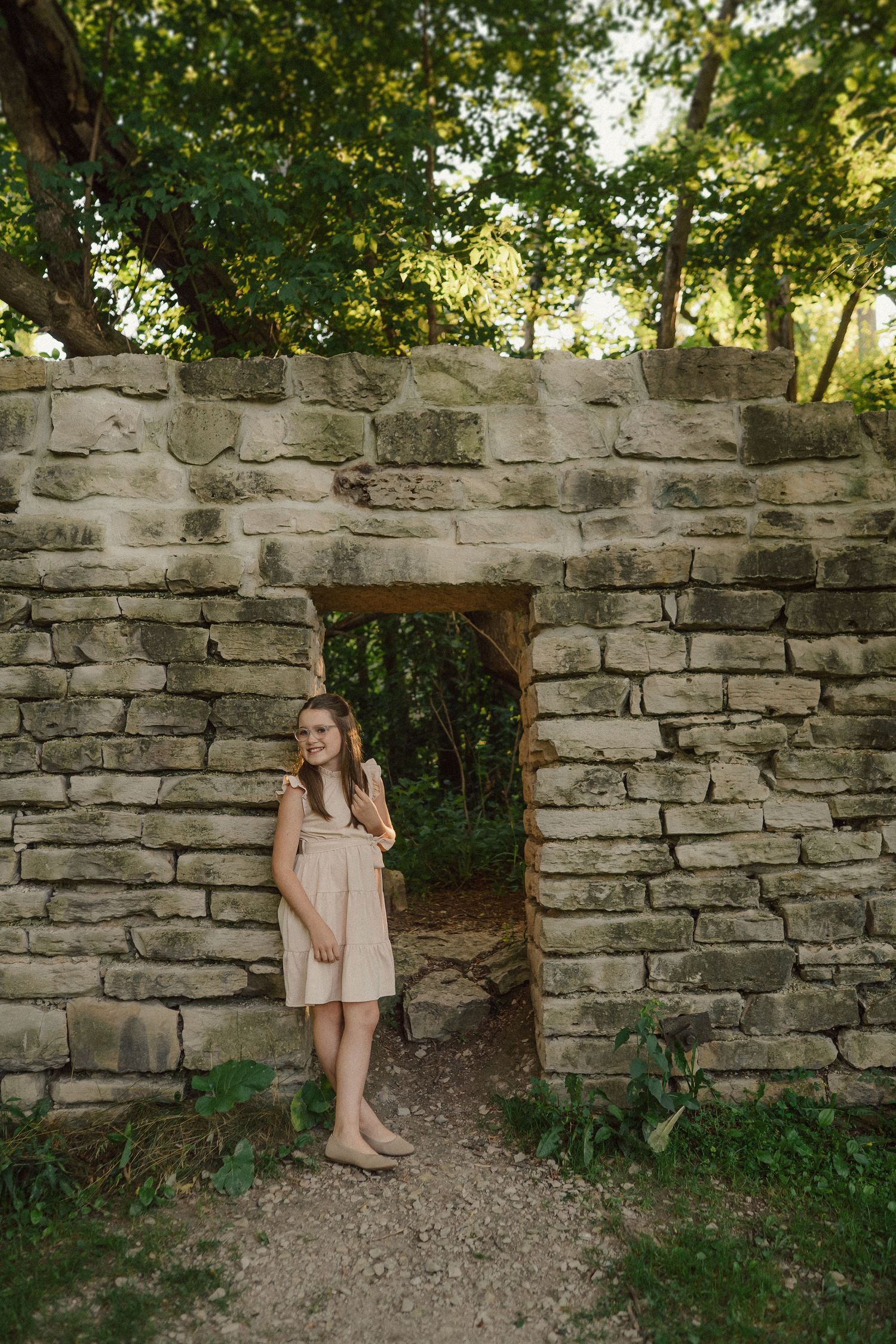 Someone in a light dress stands in a stone archway surrounded by trees and greenery in a natural outdoor setting.