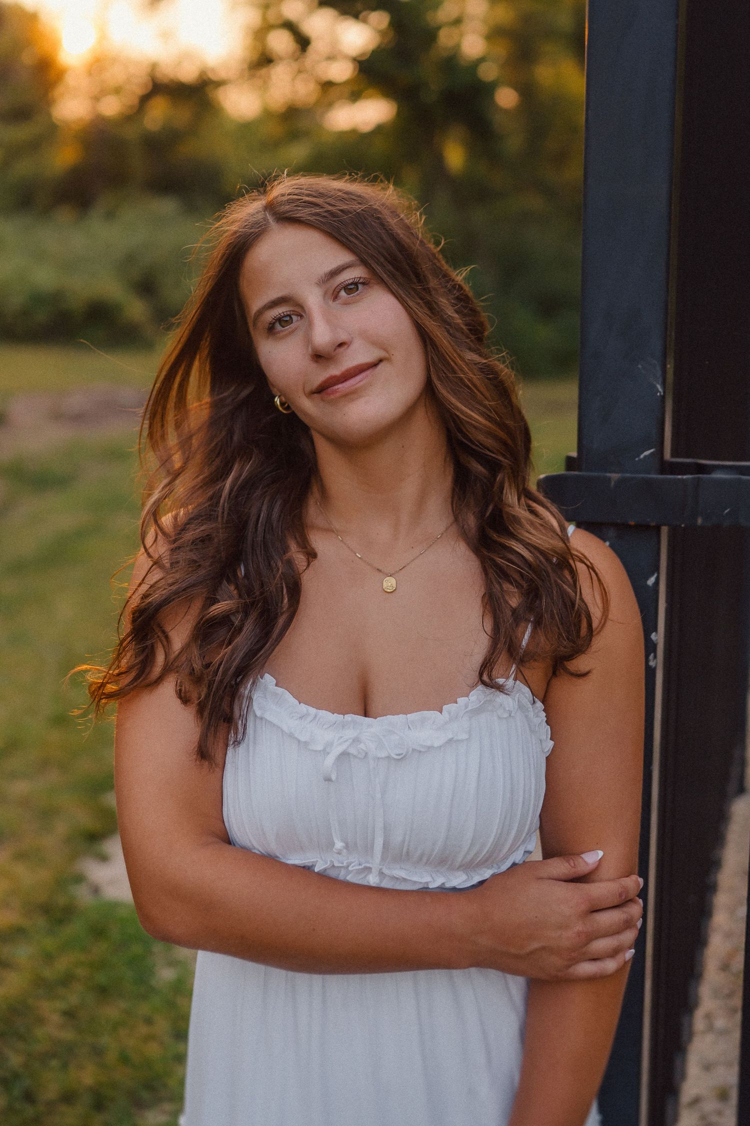 A person in white dress stands against dark post during golden hour.