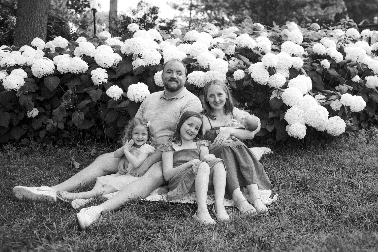 Black and white photo of family sitting by flowering bushes in natural outdoor setting.
