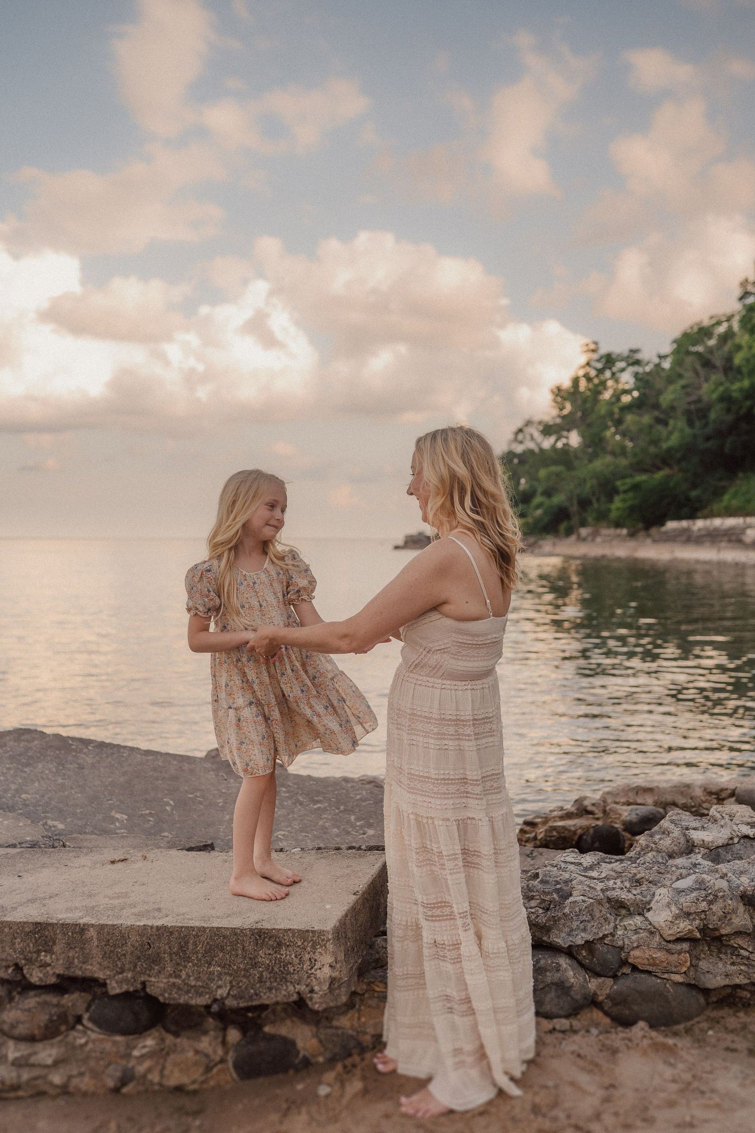 A tender moment between parent and child standing on rocky beach at sunset with calm waters and cloudy sky in background.
