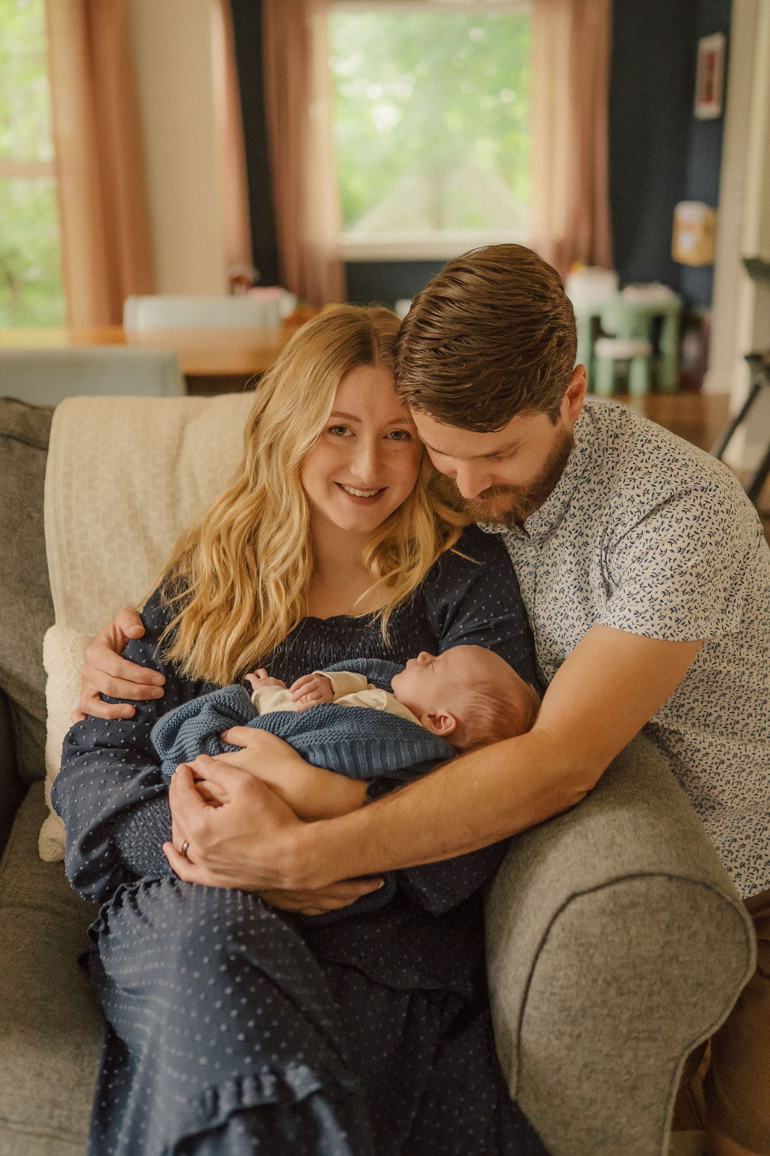 A couple cuddles with their newborn baby on a grey sofa in a sunlit living room.