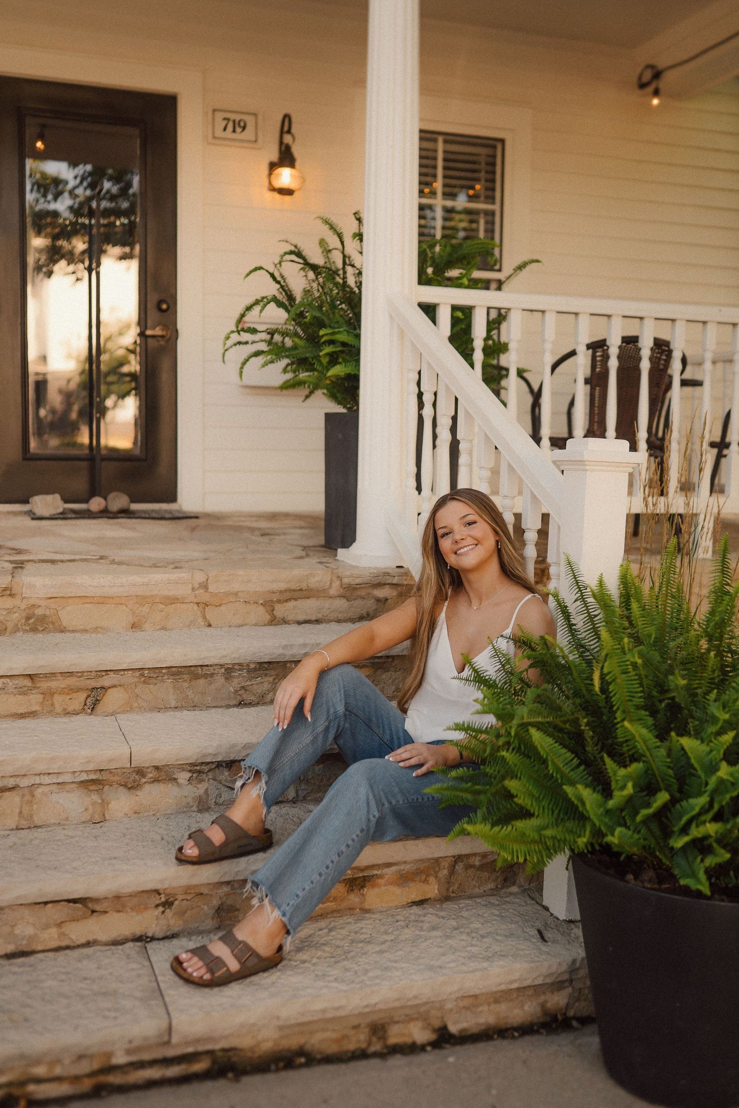 Woman sitting casually on porch steps with potted ferns and white railings during golden hour.