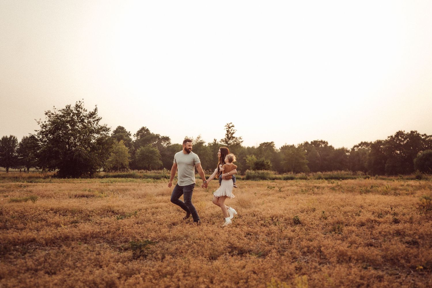 A couple walks hand in hand through a sun-drenched field at dusk.