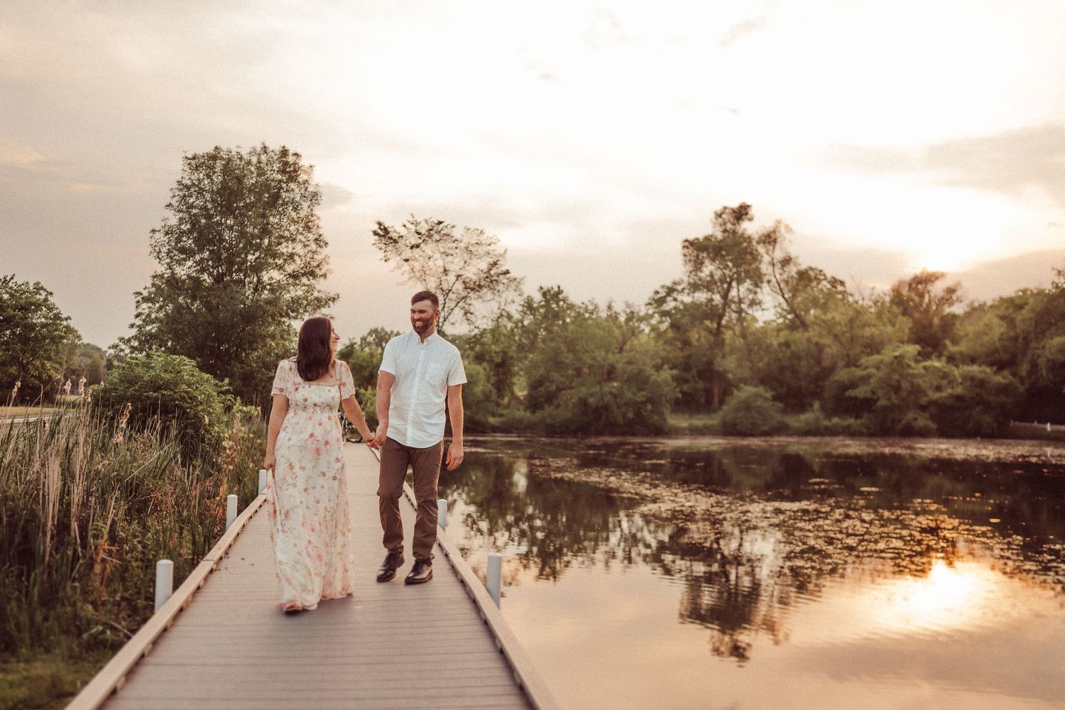 Couple strolling hand in hand on wooden boardwalk with sunset reflecting on water.
