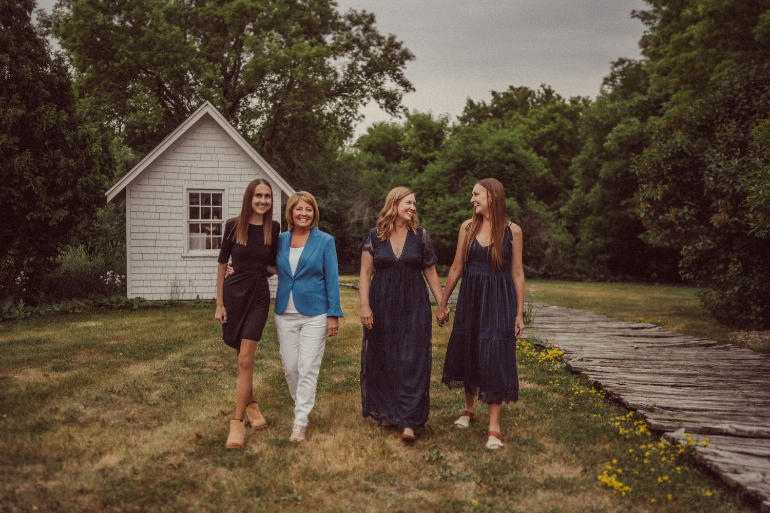Four women walking together on wooden path with white cottage in background.