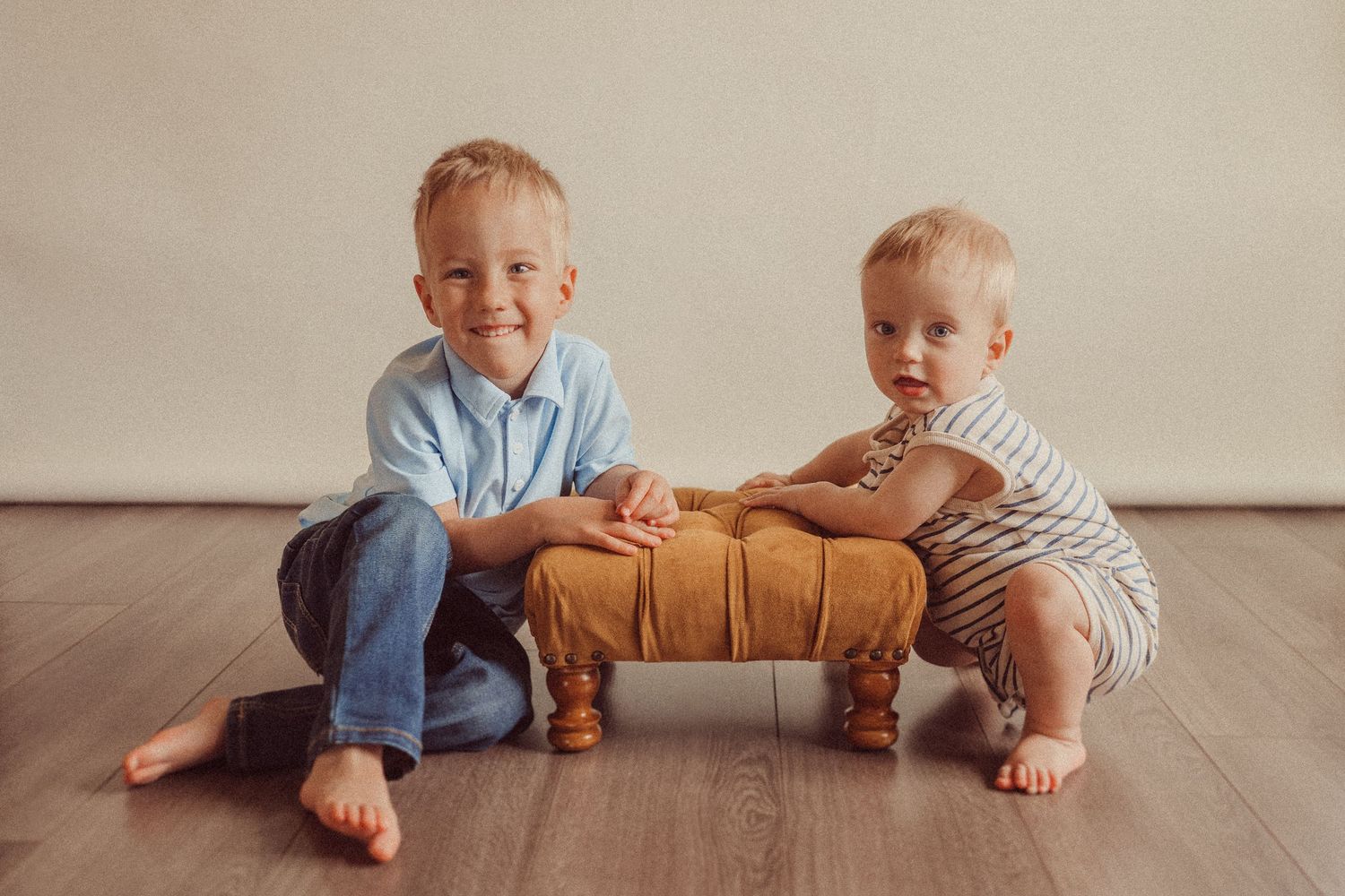 Two children posing together with wooden bench on hardwood floor indoors.