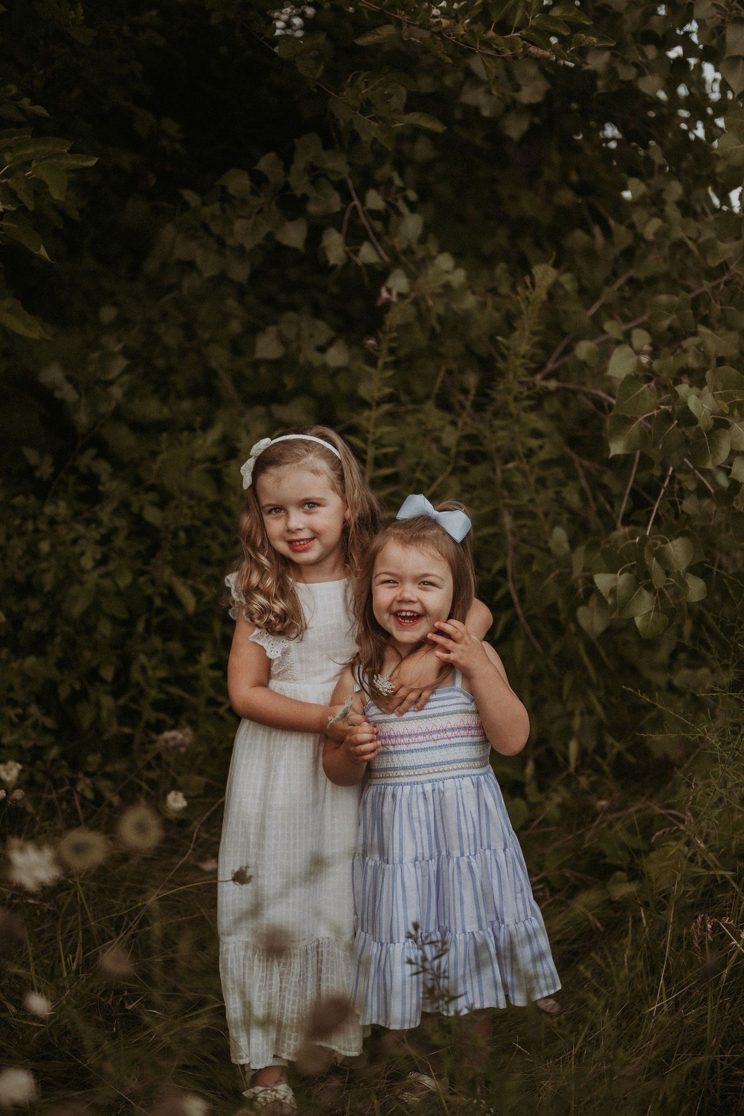 Two children in light dresses share a joyful moment together outdoors among leafy greenery.