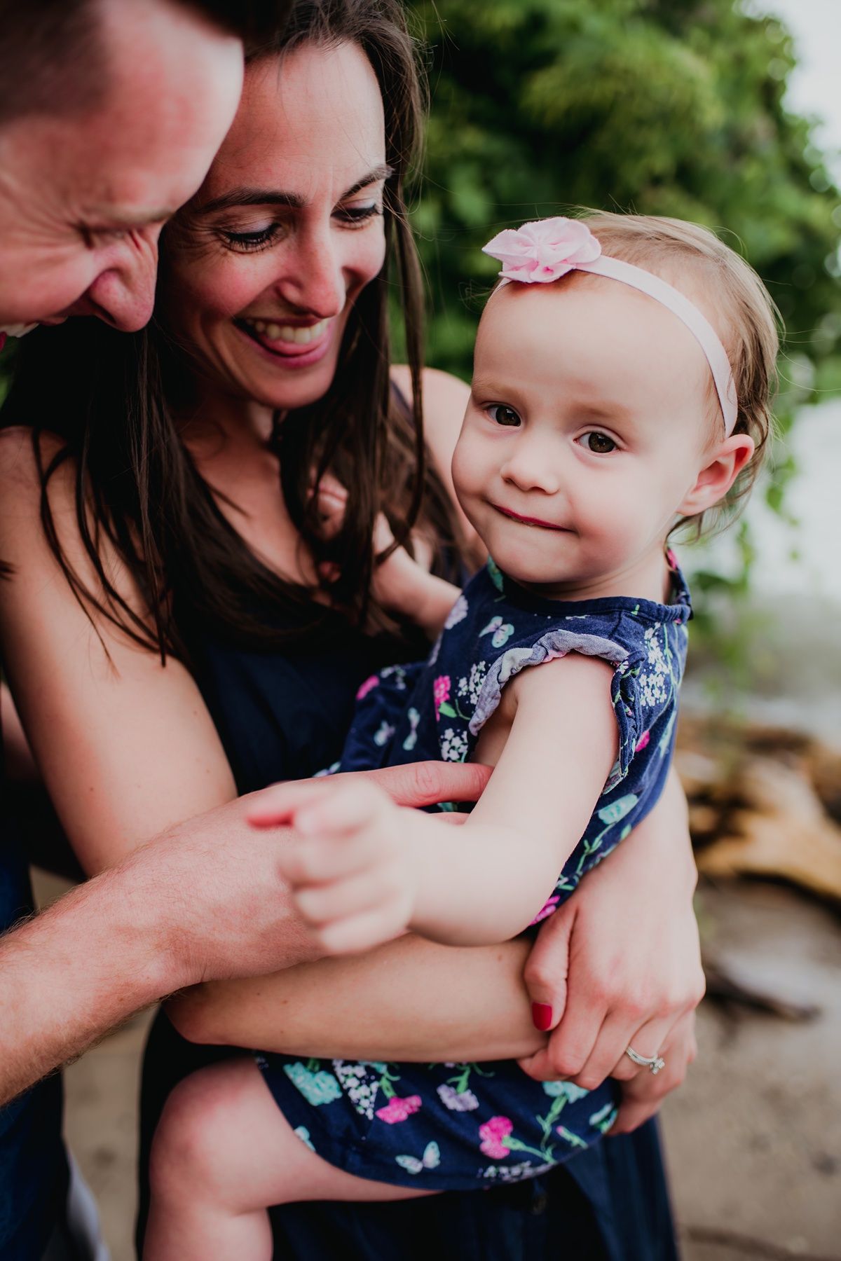 A tender family moment captured outdoors with a baby wearing a floral dress and white headband.
