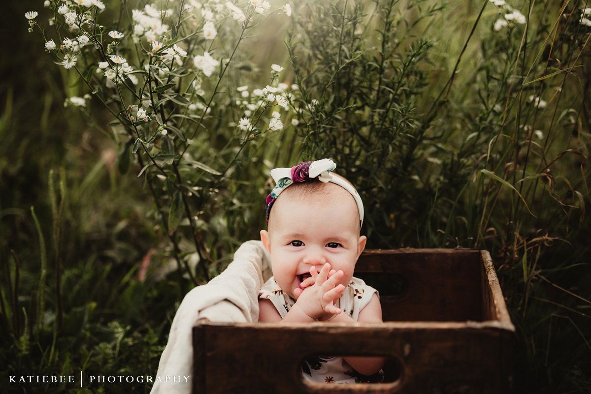 Laughing baby in white dress and bow sits in rustic wooden box among wildflowers.