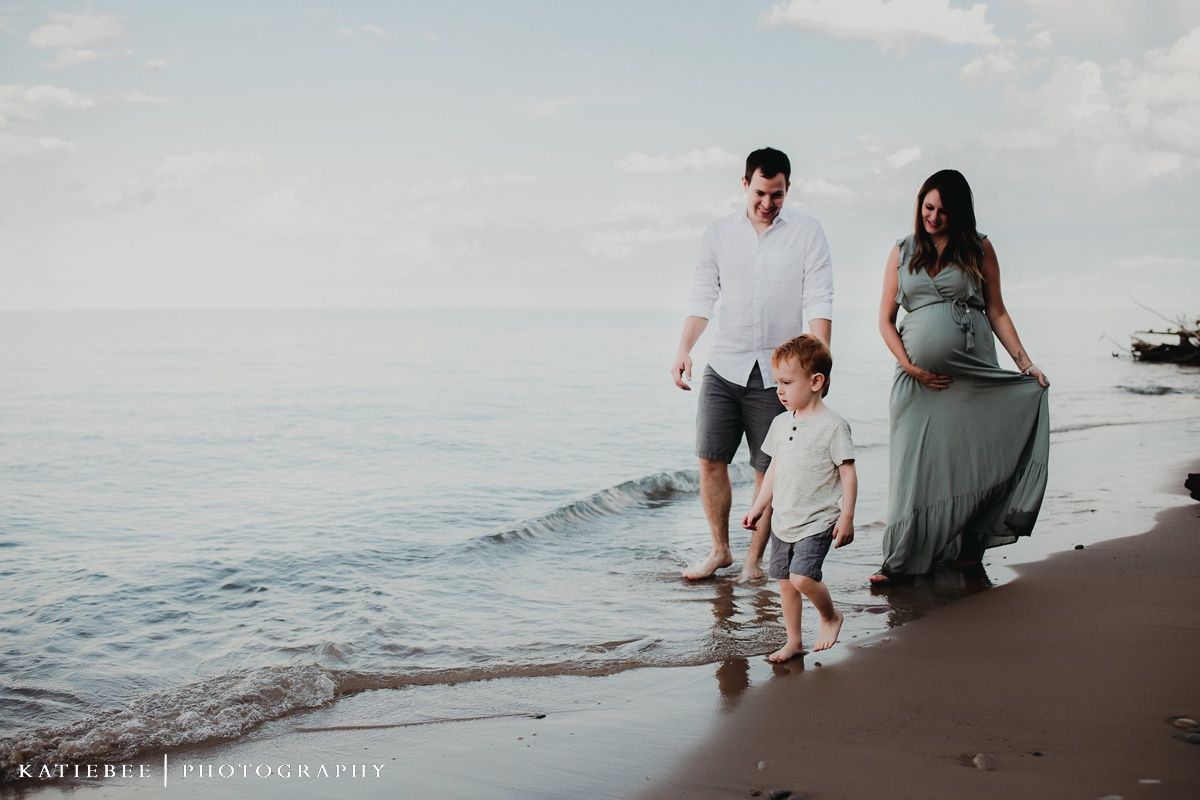 Family beach portrait with expecting mother in sage dress walking along shoreline.