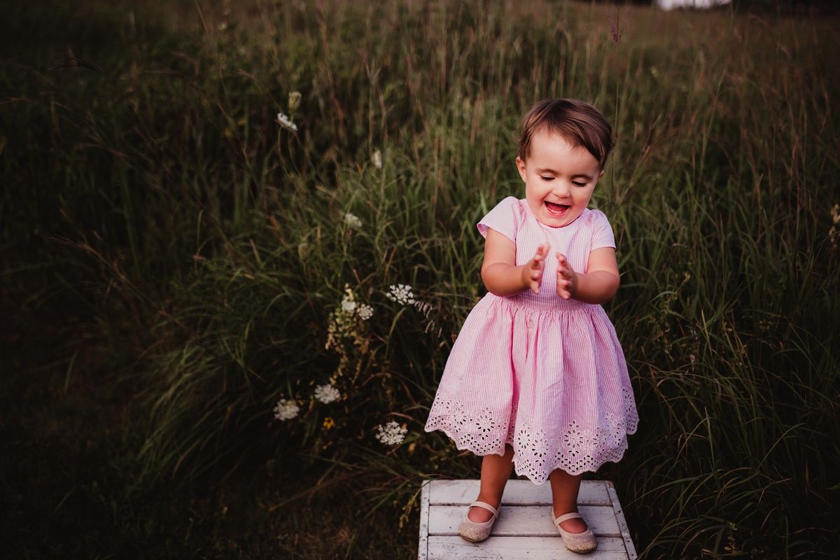 A small child in a pink dress stands on a wooden platform surrounded by tall grass.
