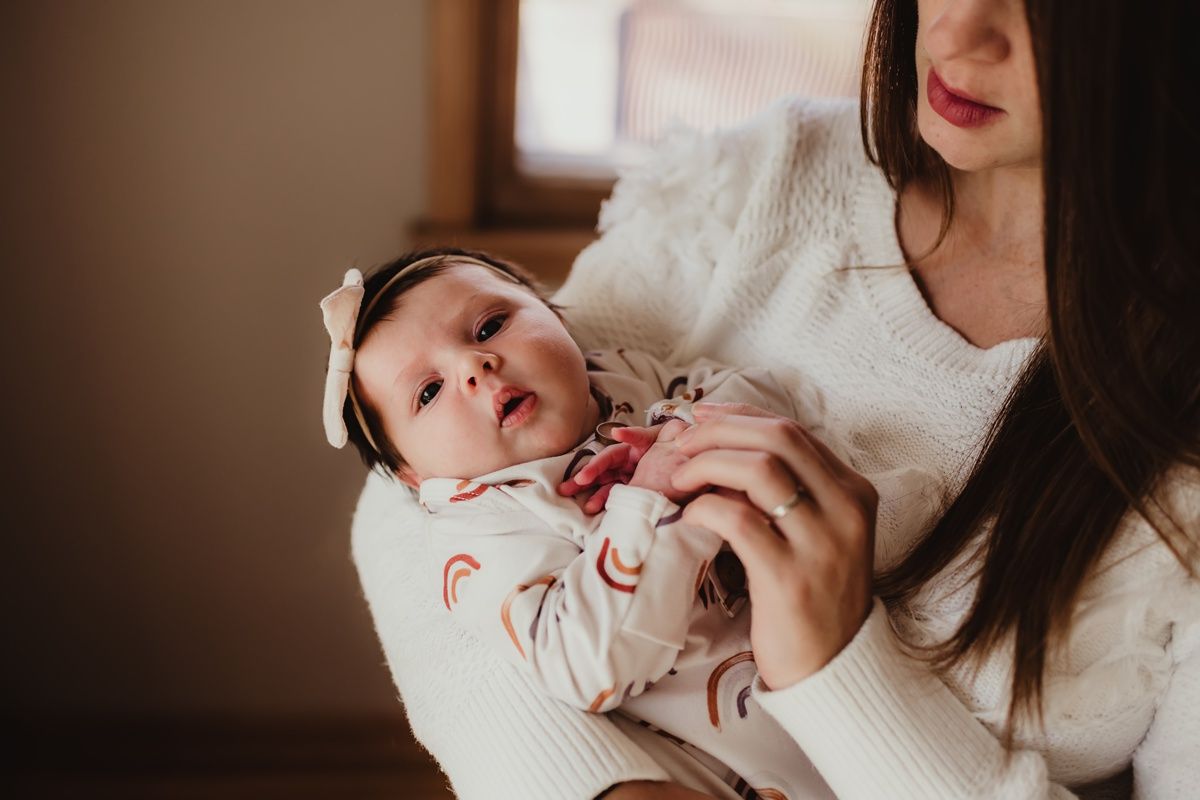 Mother standing by window holding baby in white patterned outfit.