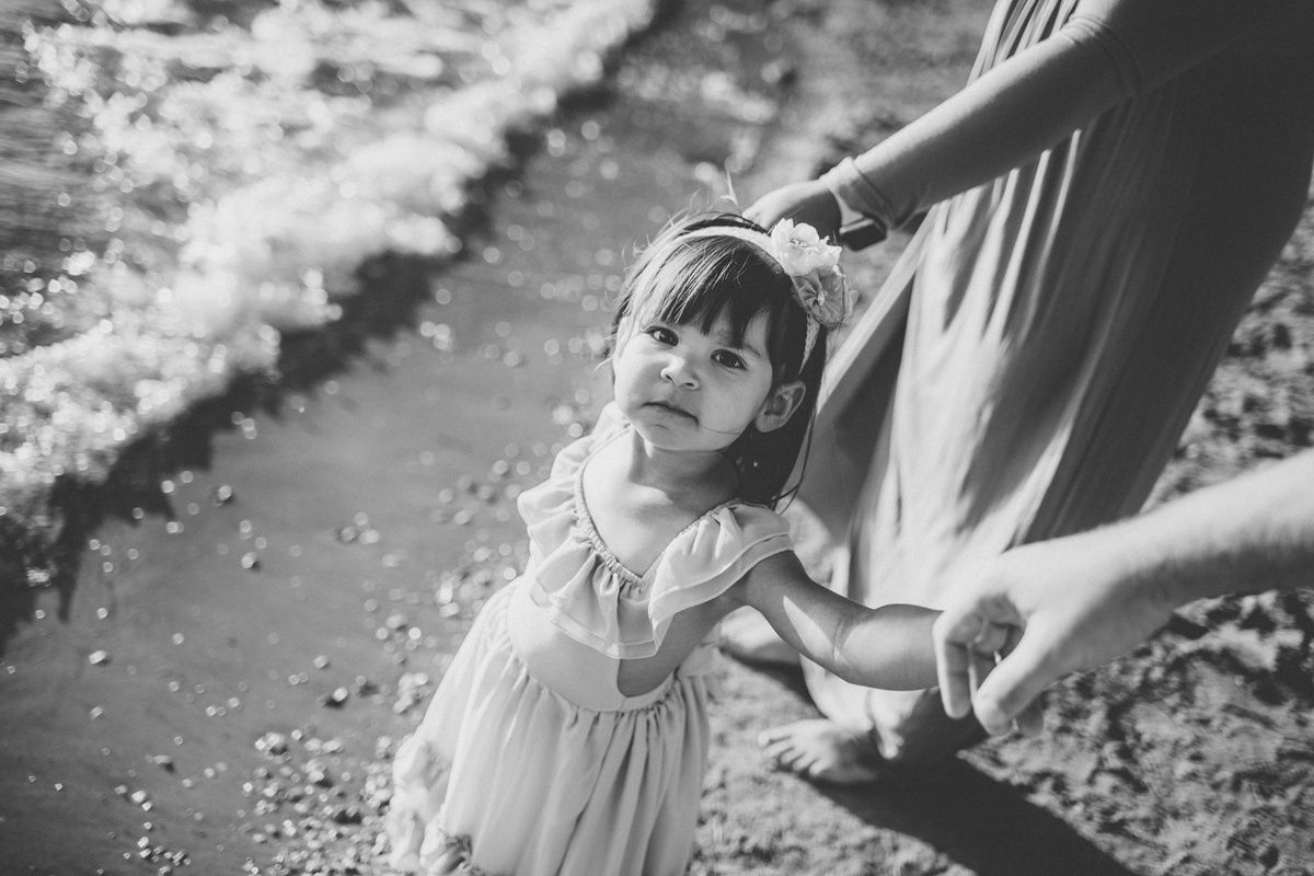 A playful black and white photo of a child in a ruffled dress dancing near water.