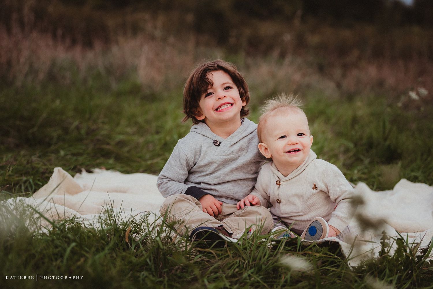Two children sit together on a white blanket in a grassy field during sunset.