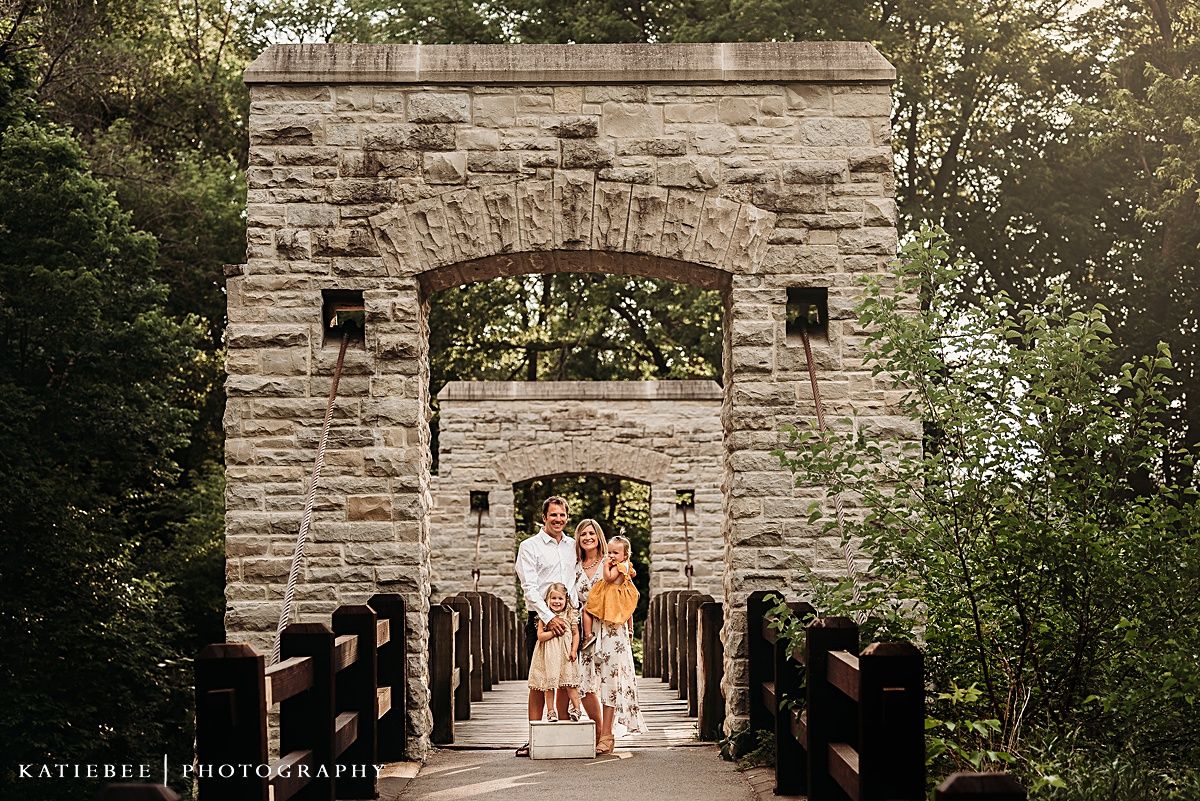 Family portrait framed by stone archway on historic bridge during golden hour.