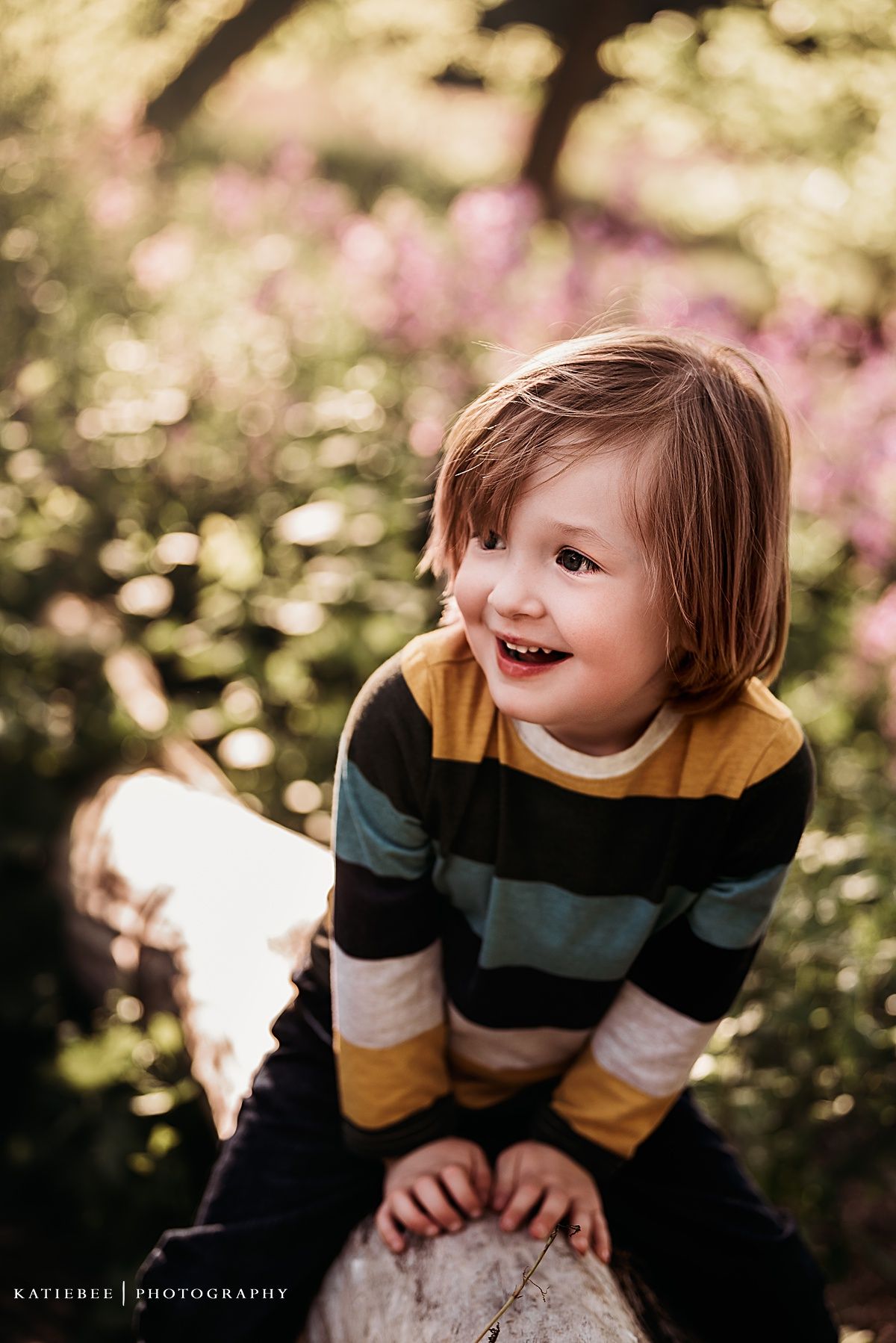 Child smiles brightly while sitting on log surrounded by bokeh and pink flowers.