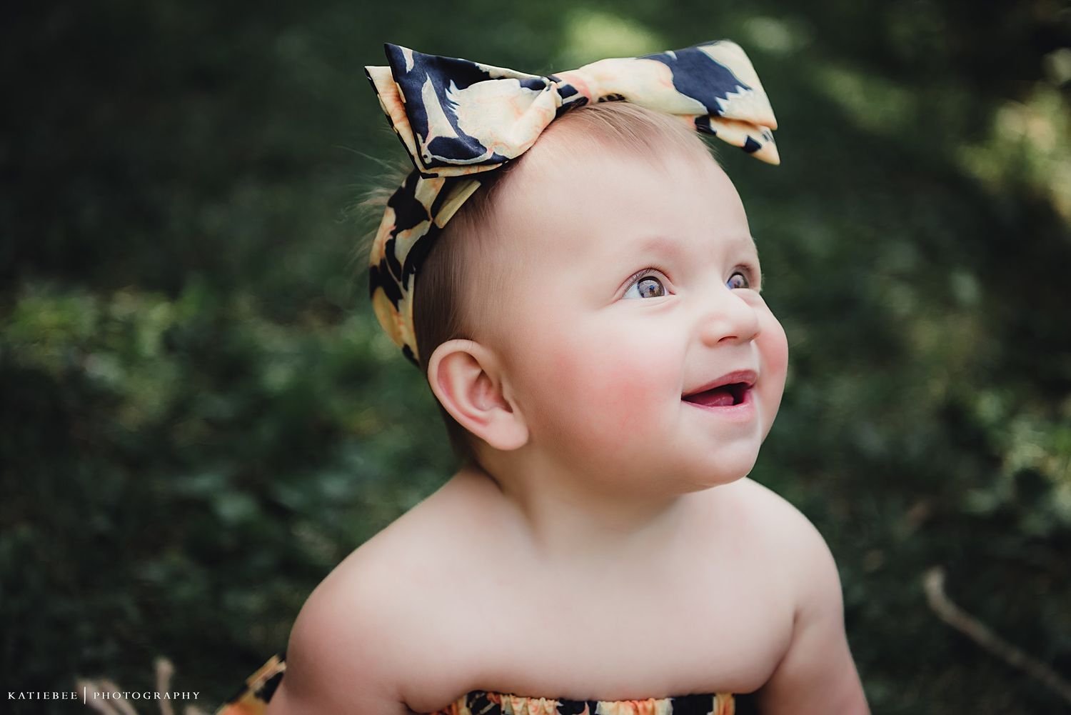 Baby in floral outfit with matching bow outdoors against green foliage.