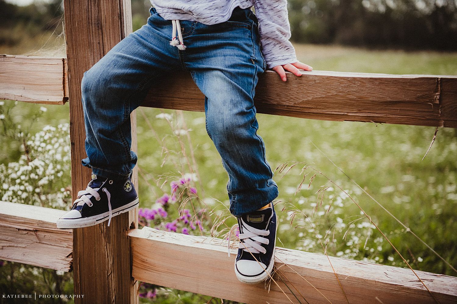 Little legs in jeans and sneakers sitting on a wooden fence with wildflowers in the background.
