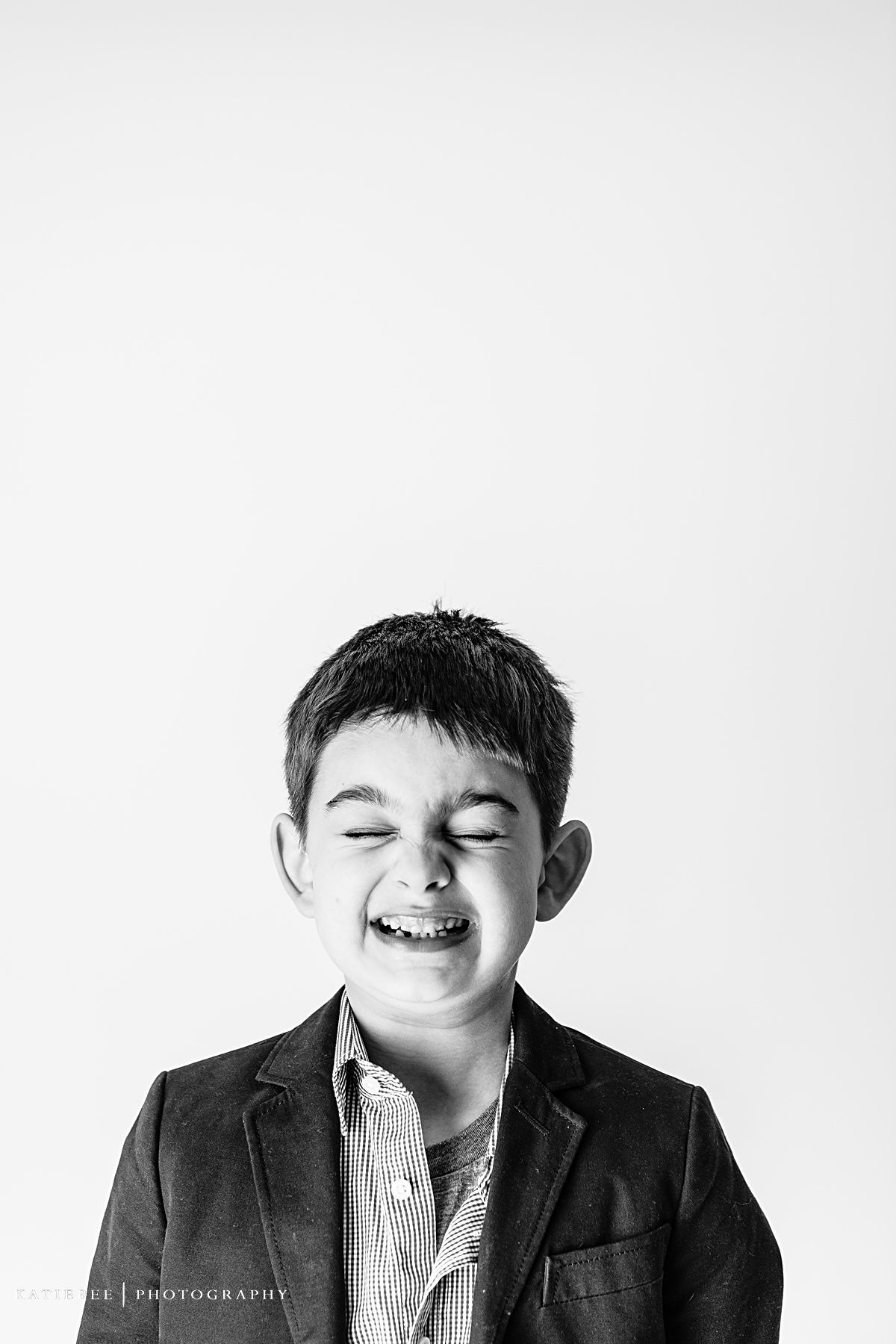 Black and white portrait of young child in blazer laughing with eyes closed against white background.