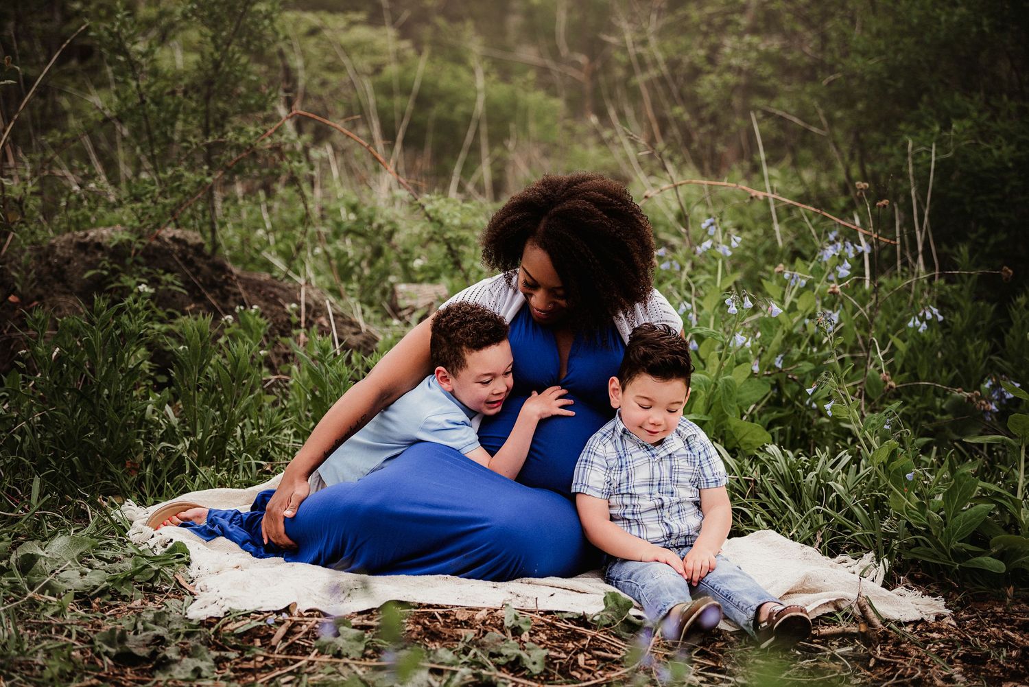 Family maternity photo session with two children on blanket amid blooming flowers.