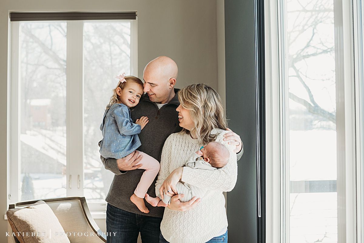 A loving family portrait with parents embracing their toddler and newborn in a sun-lit room.