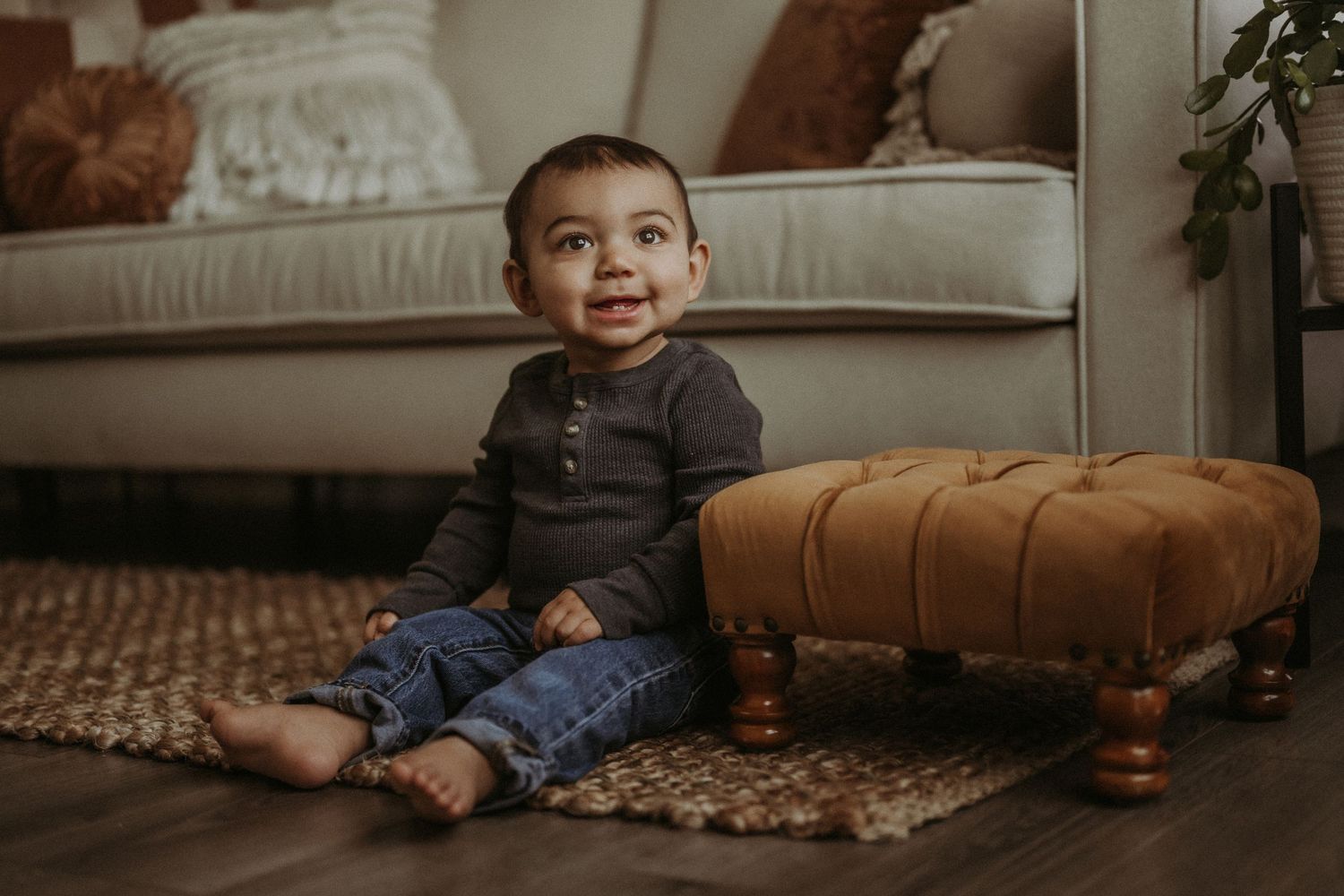 Young child in dark henley shirt and jeans sits on carpet next to brown leather ottoman.