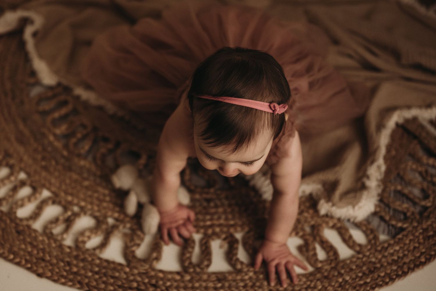 Baby wearing a headband crawls on a patterned brown rug in warm moody lighting.