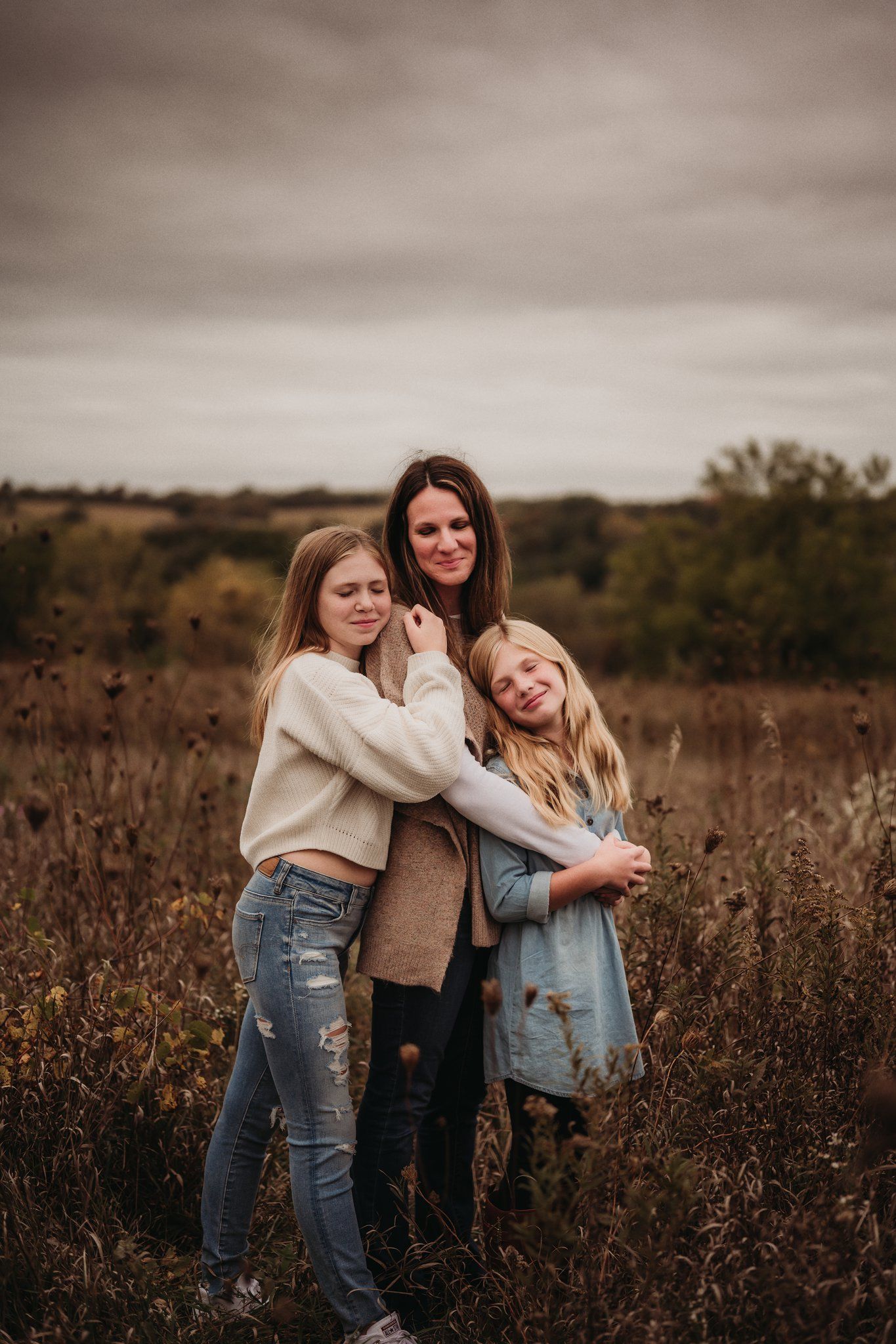 Three people embrace while standing in tall grass field during sunset.