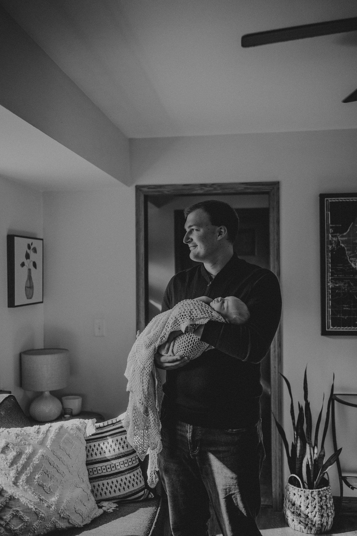 Black and white image of someone holding a baby wrapped in lace while standing in a modern living room.