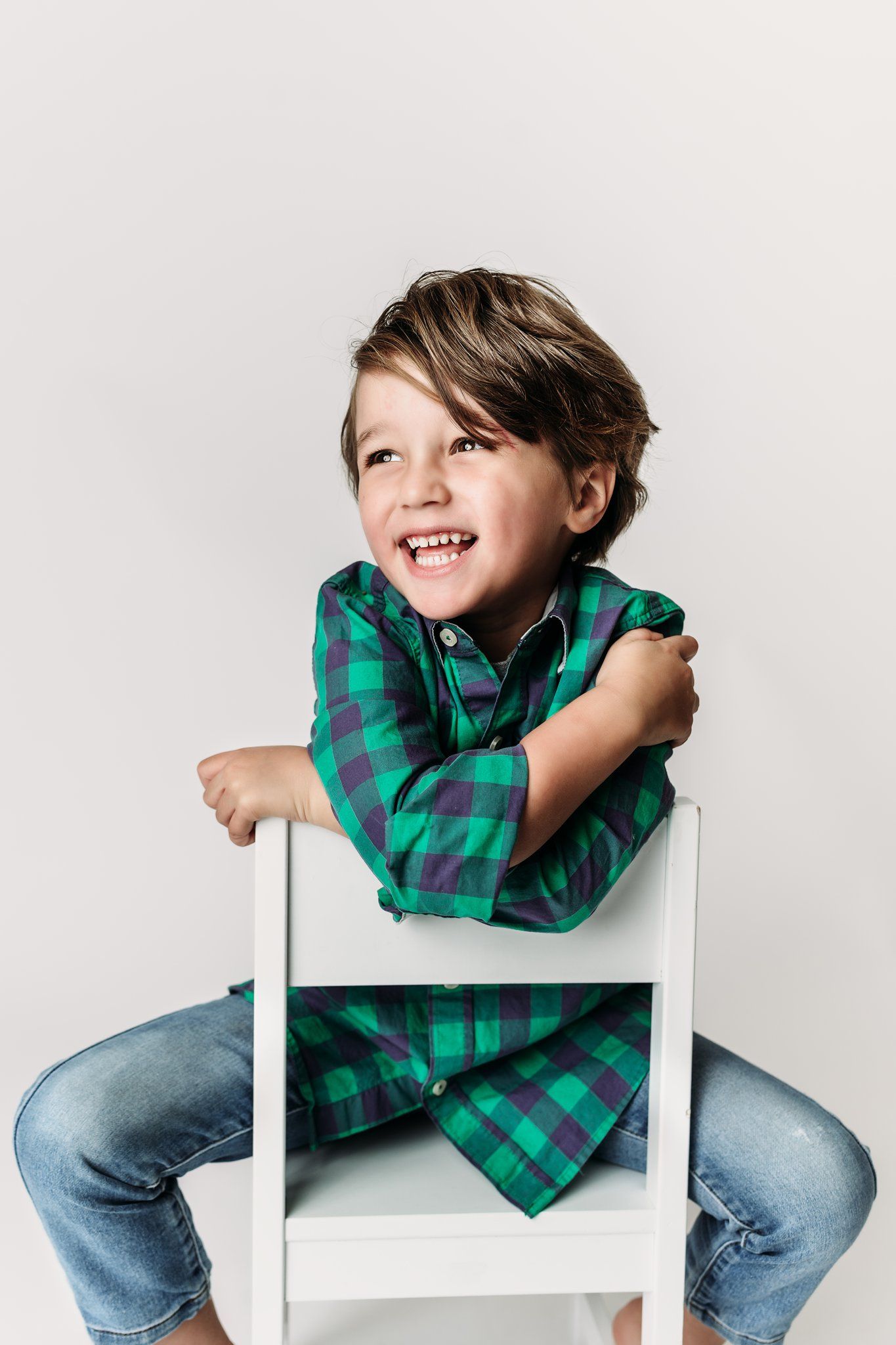 Child in green plaid shirt smiles while leaning on white chair against light background.