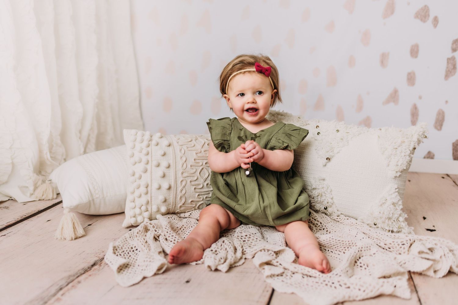 Baby in olive green dress sitting among white textured pillows.